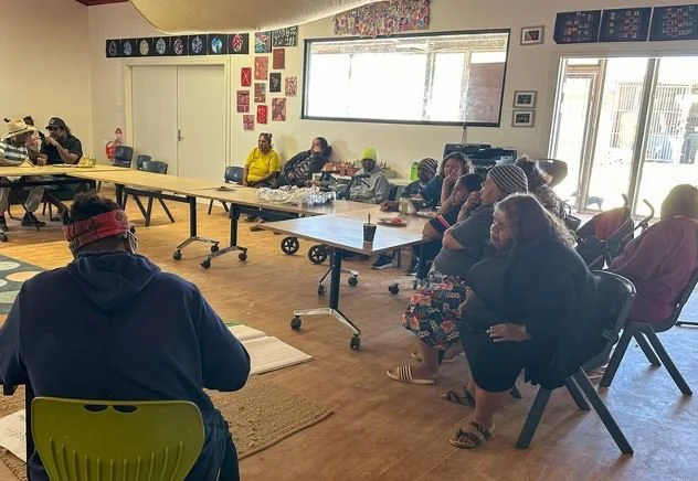 Group of people sitting in a circle in a room with windows, some taking notes, others listening attentively.