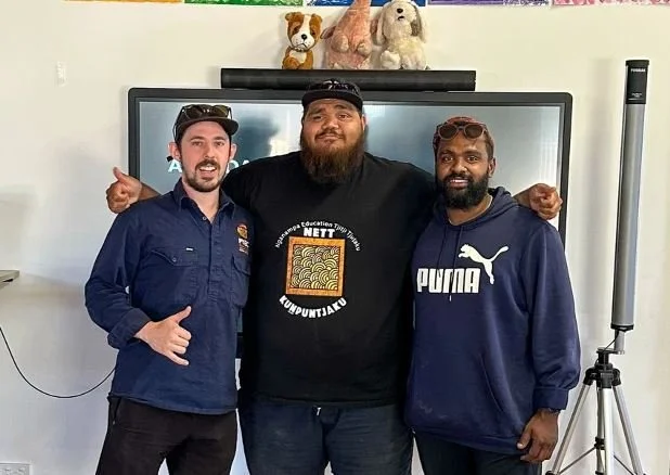 Three men standing together in front of a large screen, smiling. They are indoors, with stuffed animals on a shelf above the screen and a tripod on one side.