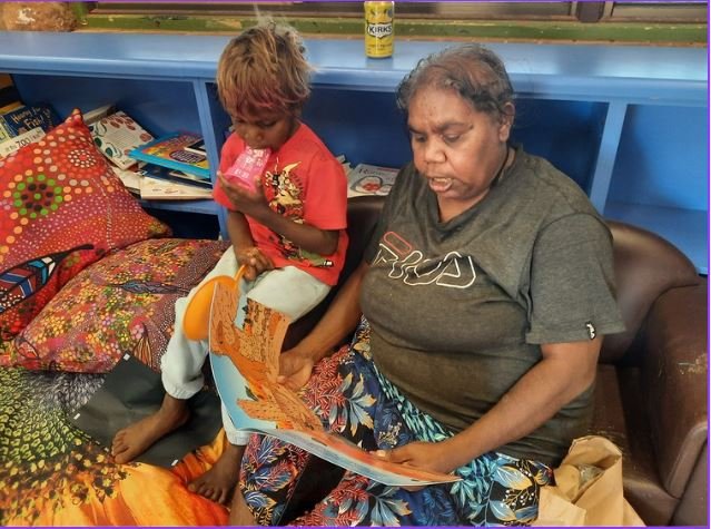 A woman and a young boy sit on a bed with colorful bedding and pillows. The woman is reading a newspaper, and the boy is holding a pink cup, looking at the newspaper. A blue shelf with books and a yellow can are behind them.