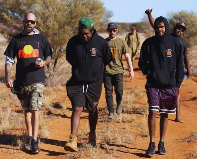 Five young men walking outdoors on a dirt trail in a desert-like environment with sparse vegetation, dressed casually with some wearing hoodies, t-shirts, and shorts, one of them holding a water bottle and another raising his fist.