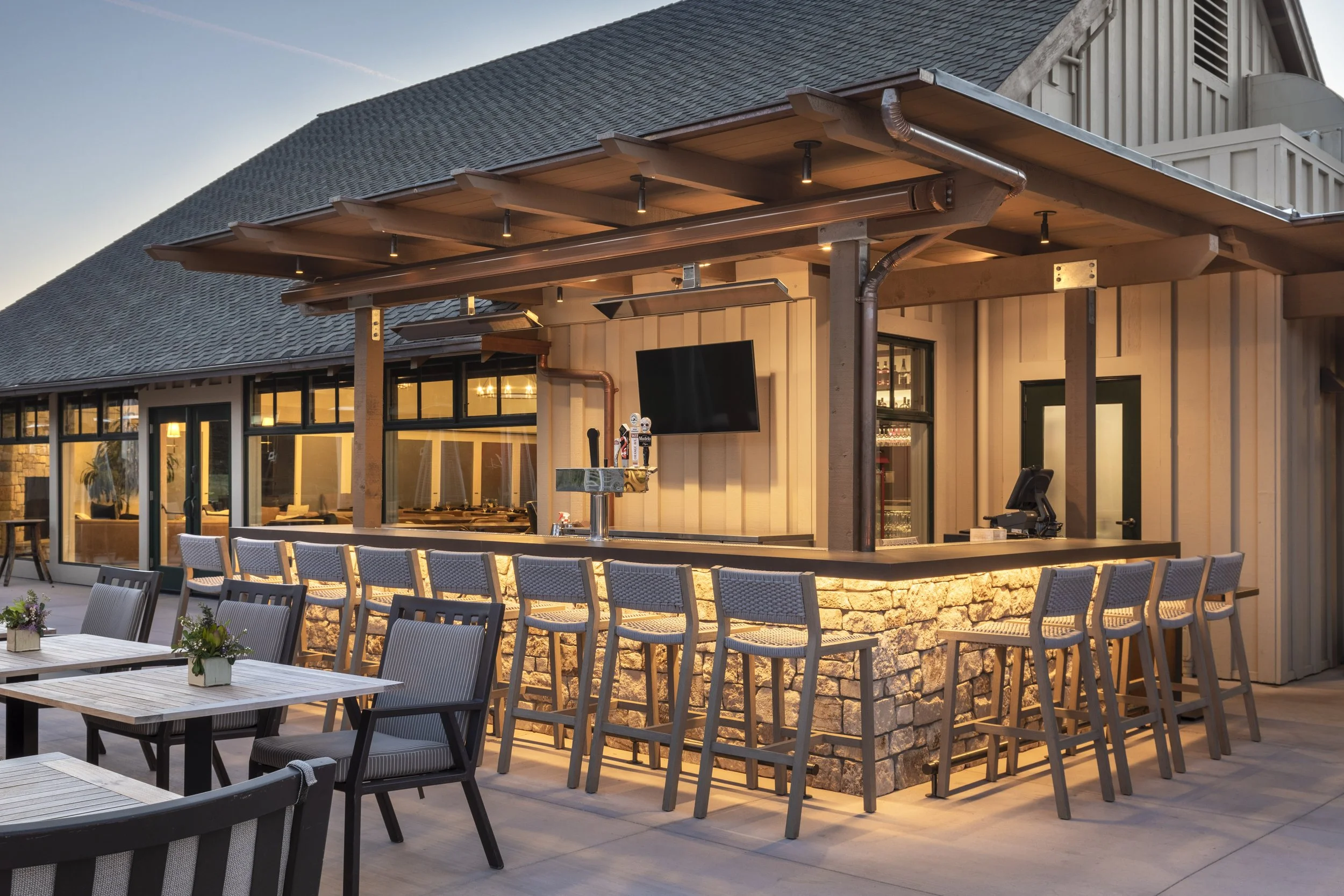 An outdoor stone-cladded bar illuminated by up-lighting, with a stone countertop, bar stools, and a TV on the wall.