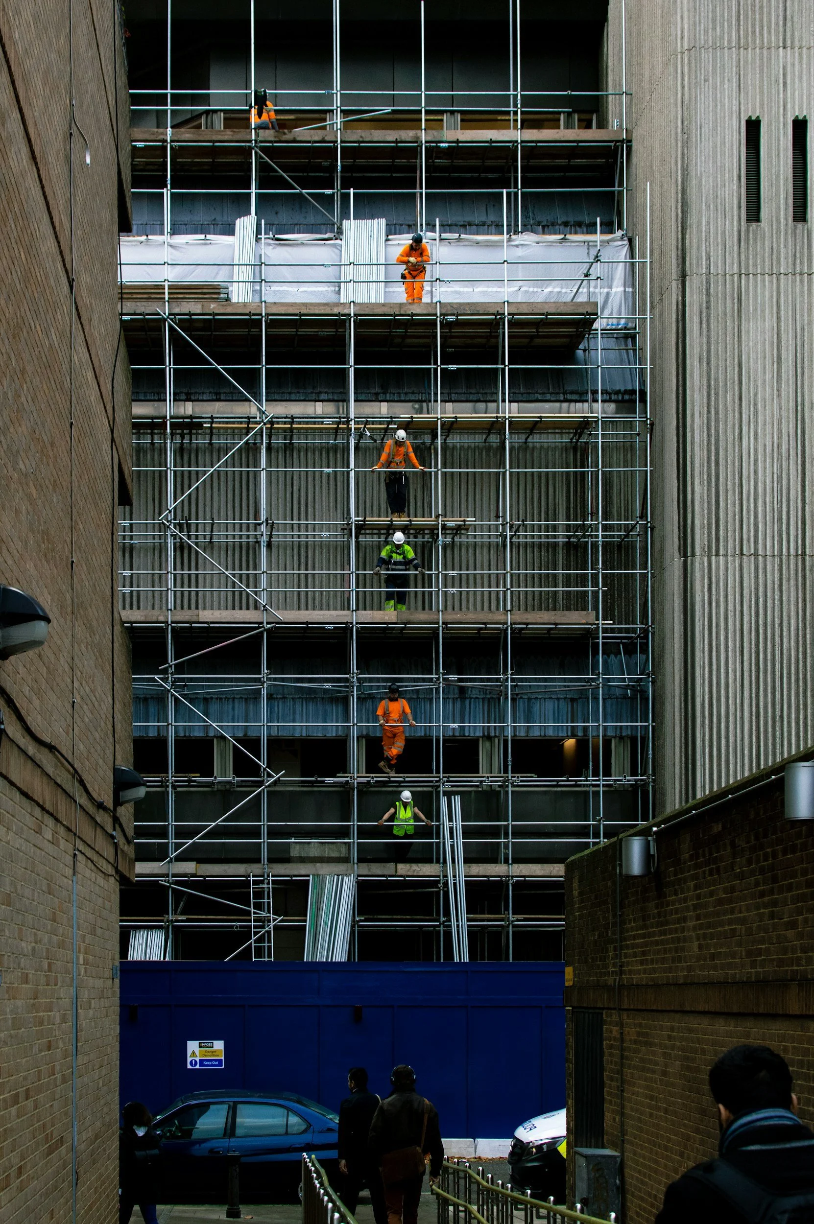 Construction workers wearing safety helmets and reflective vests working on scaffolding on a building facade, with pedestrians walking nearby.