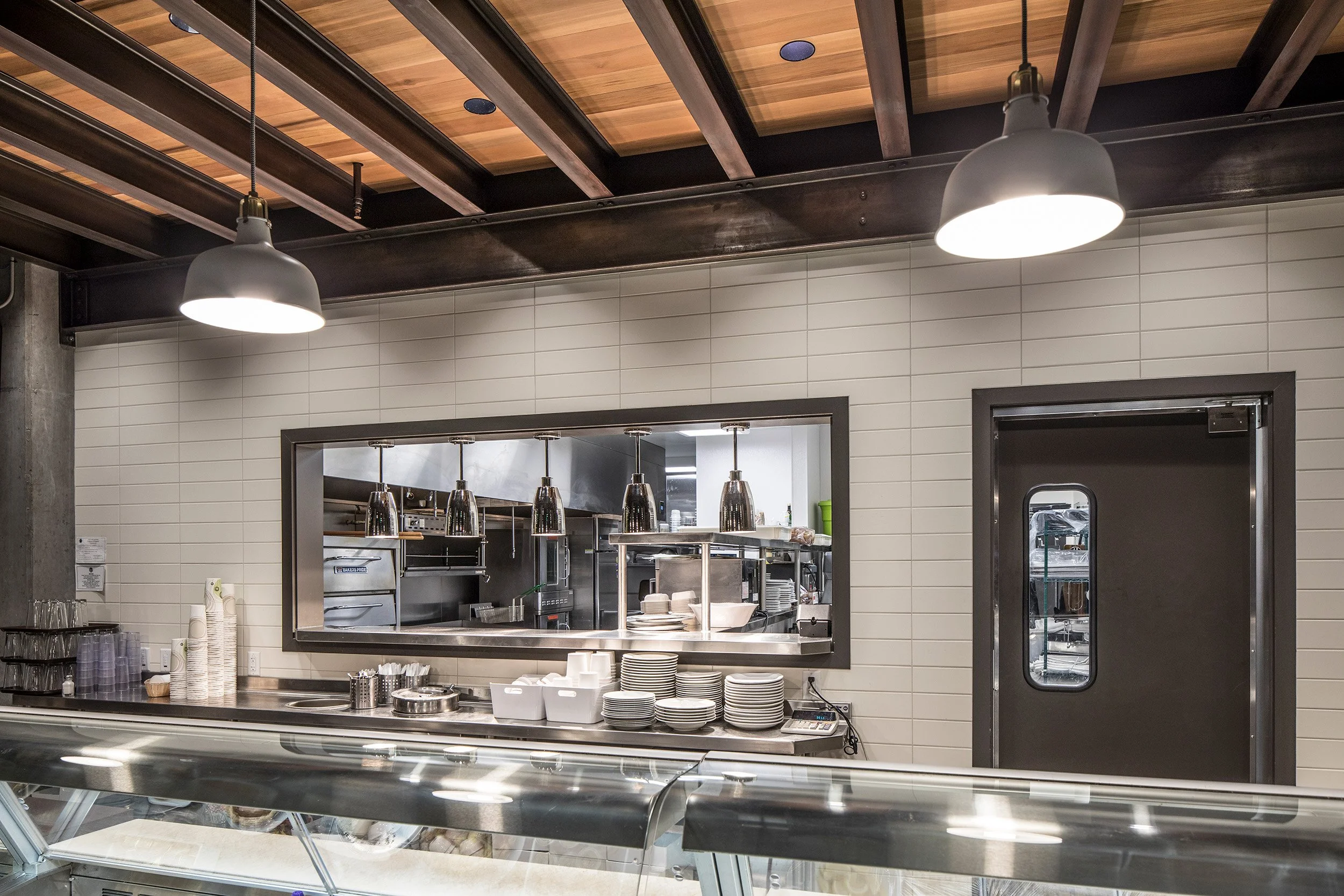 A view of a commercial kitchen through a service window, with white subway tile walls and exposed wood ceiling beams. Gray pendant lights illuminate the serving counter in the foreground.