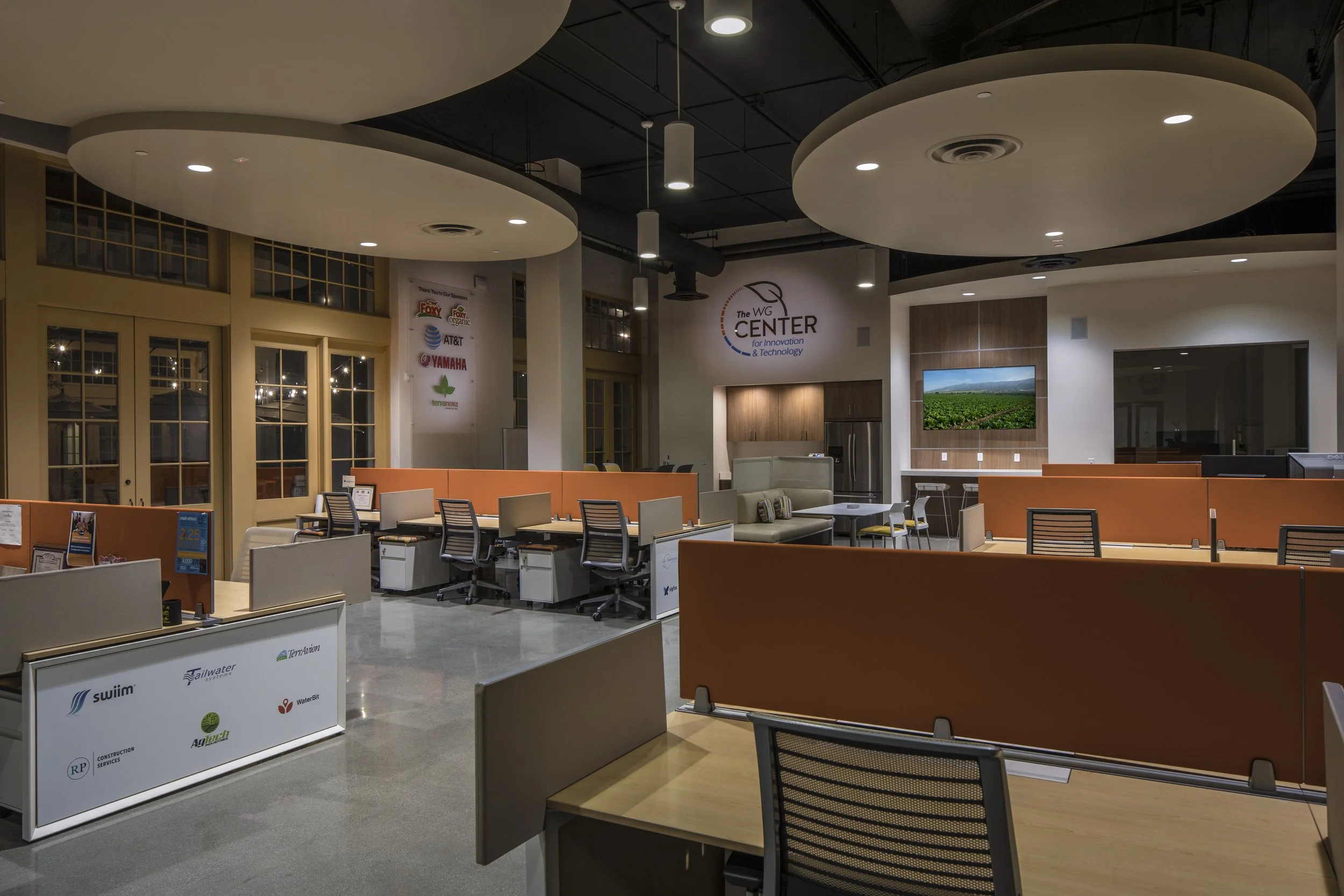 An open office featuring workstations, a mix of causal and formal seating. Polished concrete and modern lighting with cloud ceilings.