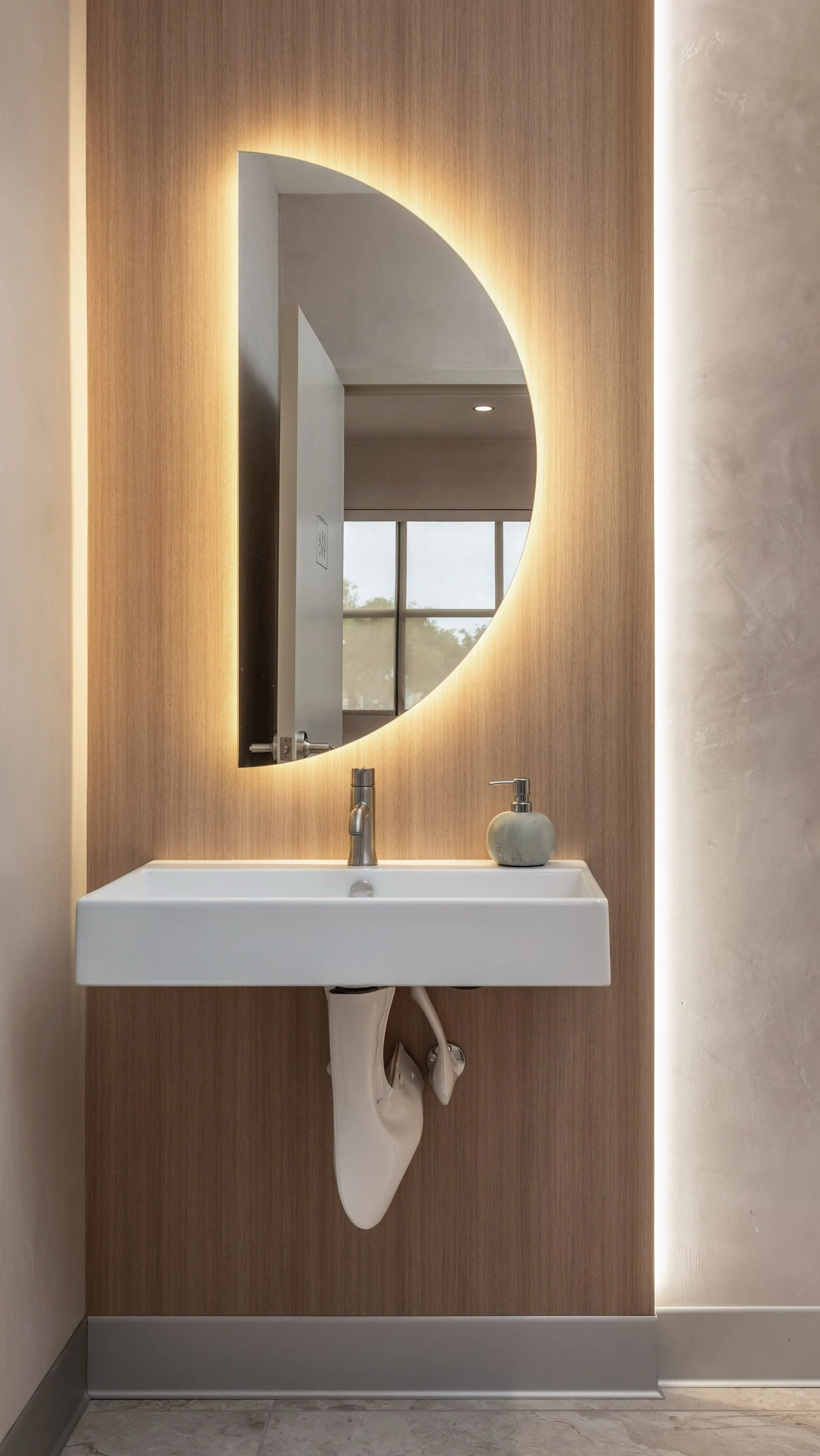 Backlit Restroom moon shaped mirror, wood laminate with edge lighting, modern design aesthetic, white pedestal sink. Gallery Aesthetics interior.
