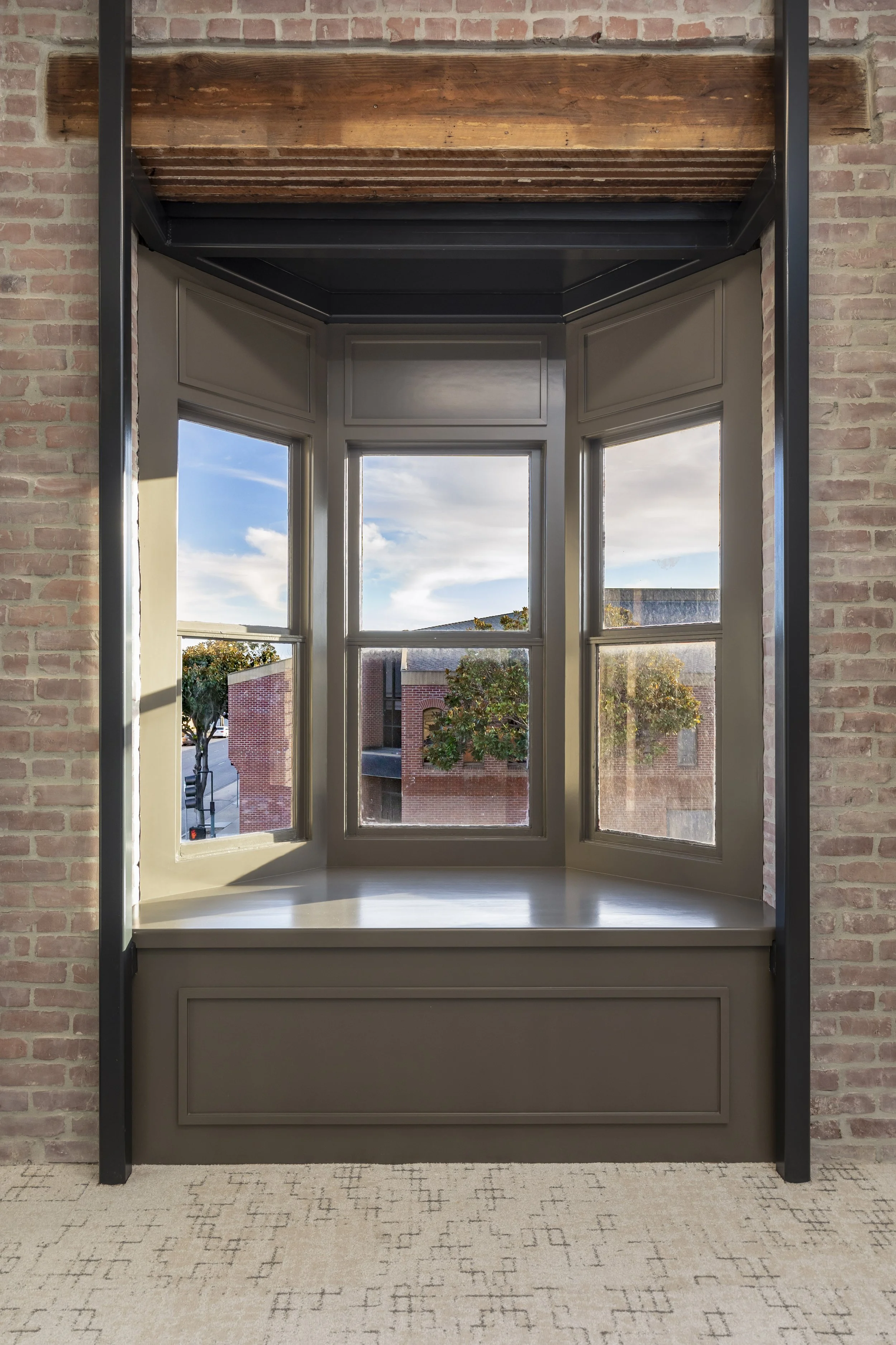 A historic bay window along an original brick wall, with a view of the city below. The window features an exposed wood header, and is flanked by metal structural posts.