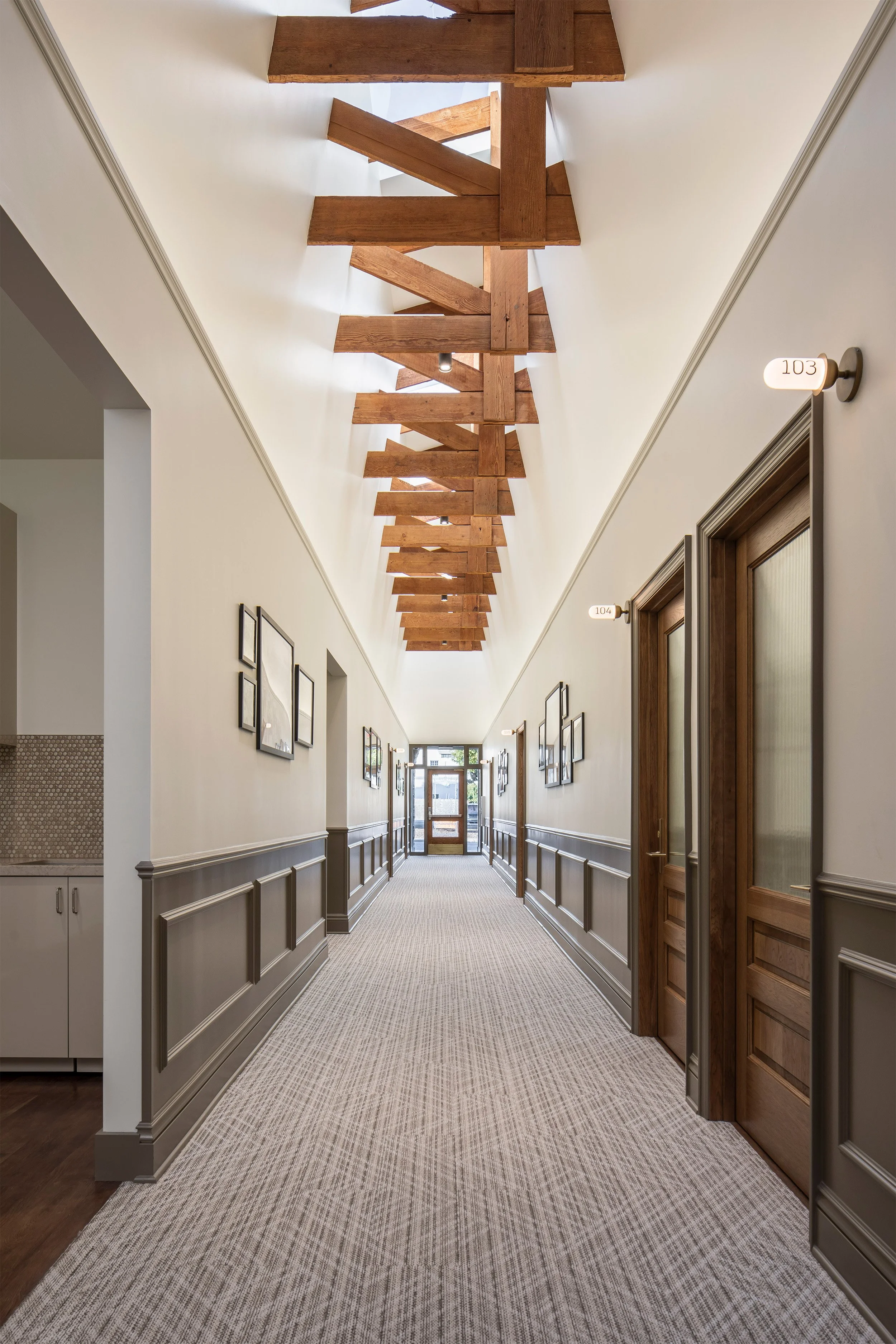 A long hallway with a patterned carpet, wooden doors, and a striking wooden beam ceiling. The hallway is lined with artwork and has a sense of historical charm.