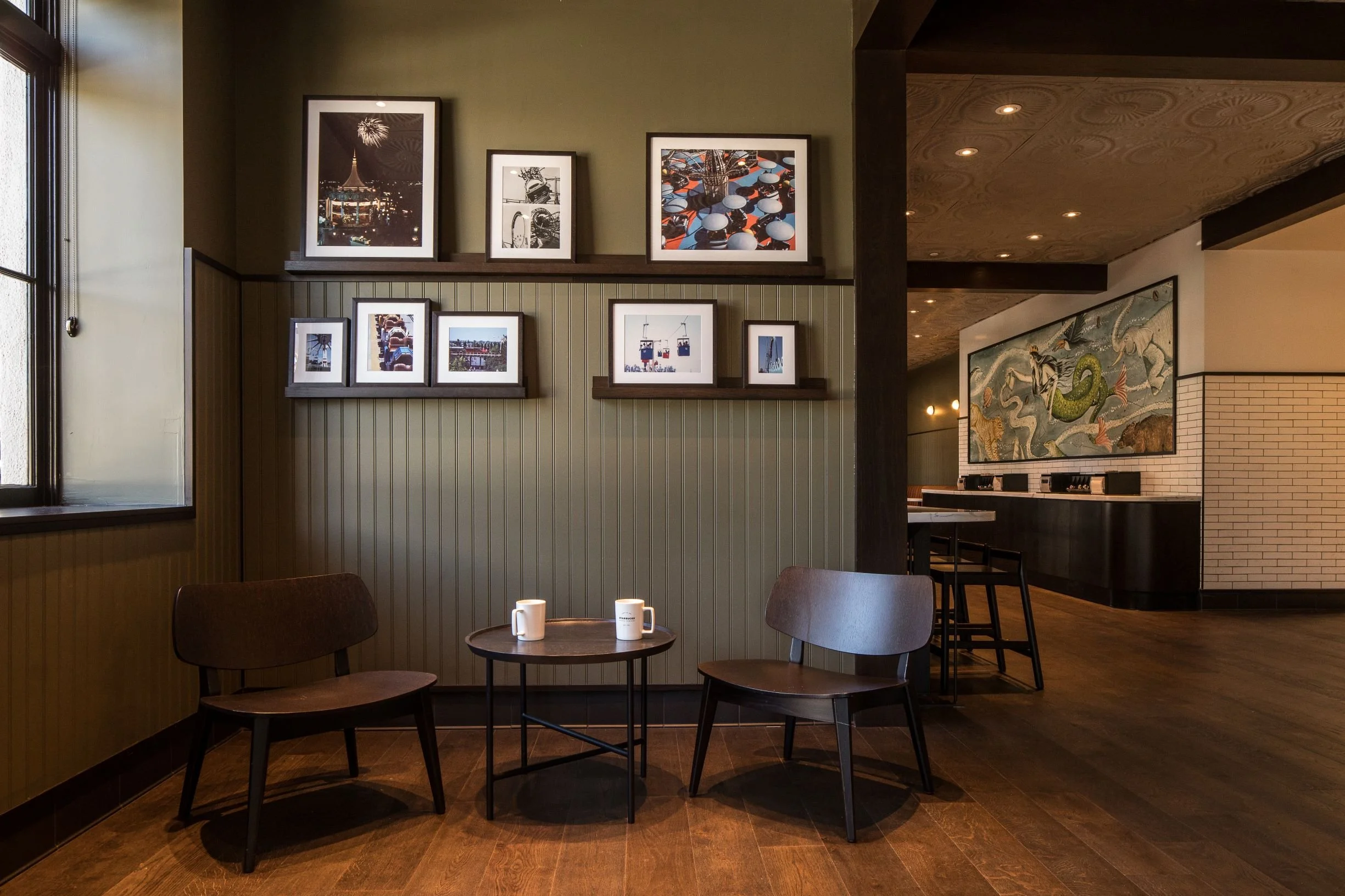 A cozy seating area in a coffee shop with two chairs and a small table. The wall behind the seating area is decorated with framed photographs. This space leads to a larger area with a counter and artwork.