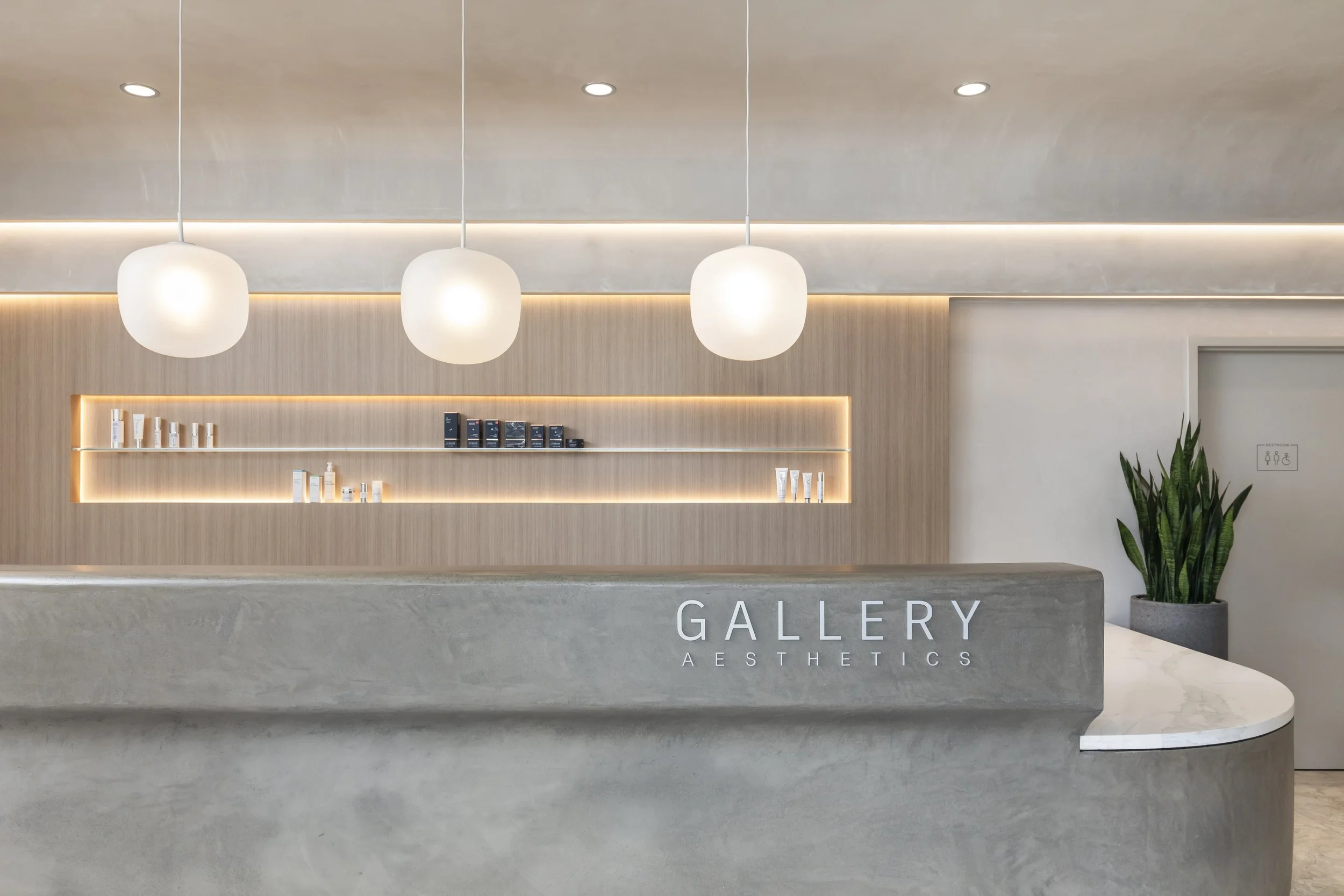 Modern reception area of a plastic surgery office, pendant lights, backlit wood shelving with skincare products, and a potted snake plant. Gallery Aesthetics interior.