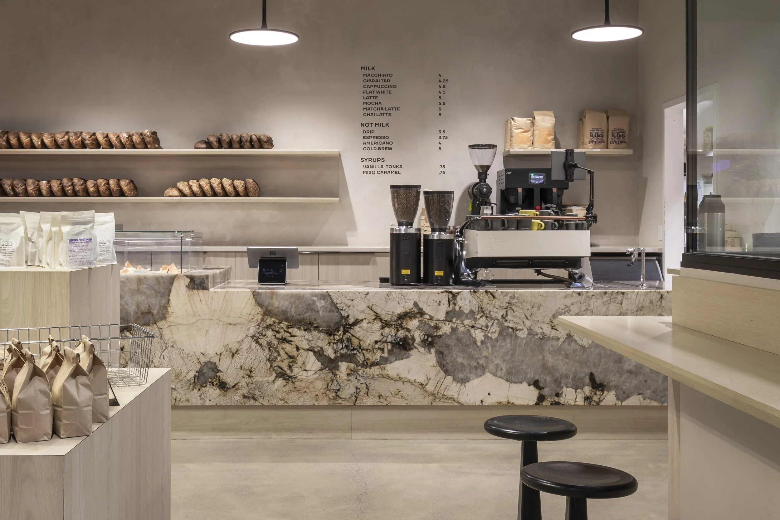 The interior of a bakery with a modern and minimalist design. A long marble countertop, shelves displaying baked goods, and an espresso machine.