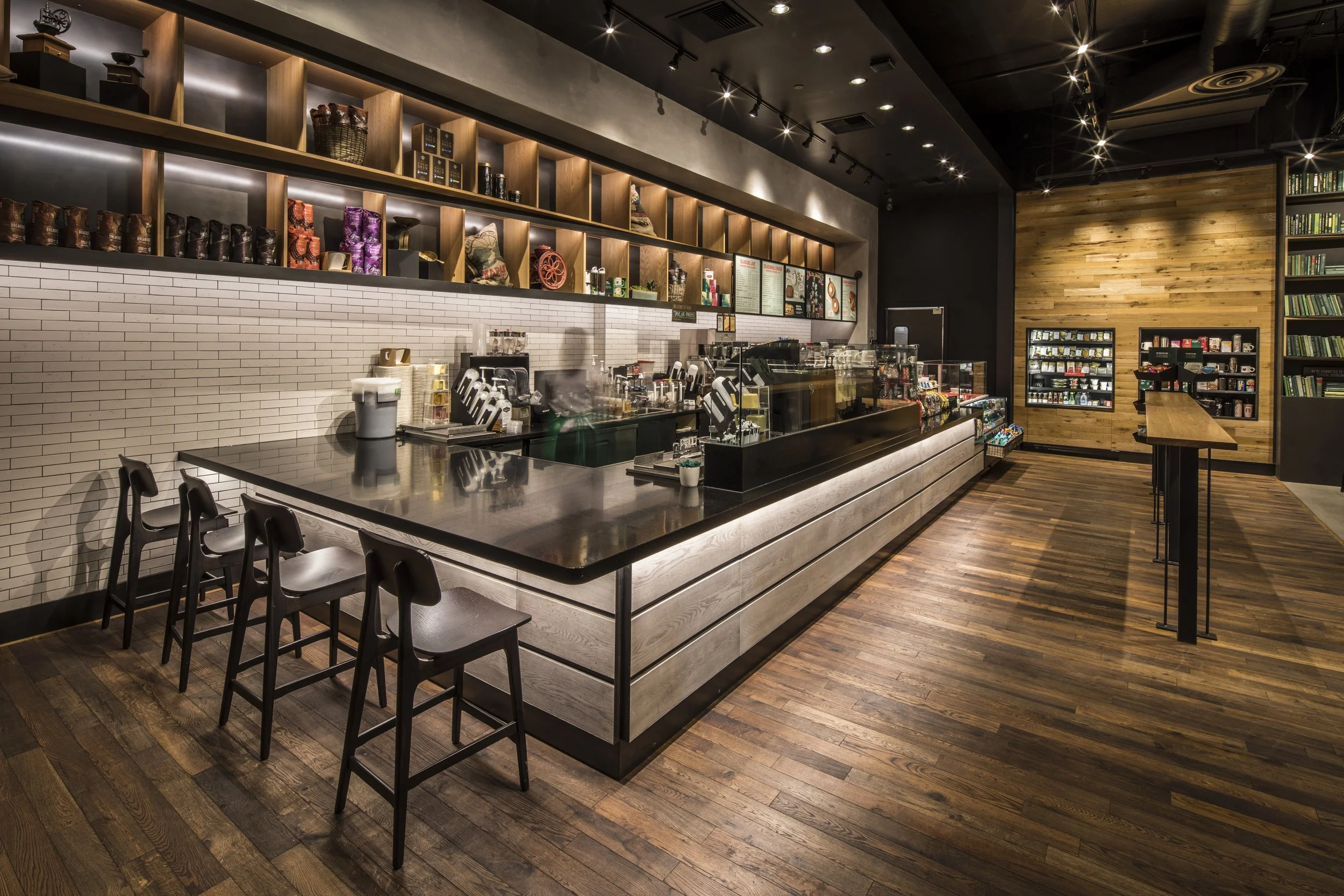 A modern coffee shop interior with a long, dark countertop, white brick backsplash, and wooden shelves filled with decorative items. Four black chairs are tucked under the counter, and warm lighting highlights the polished wood floor.