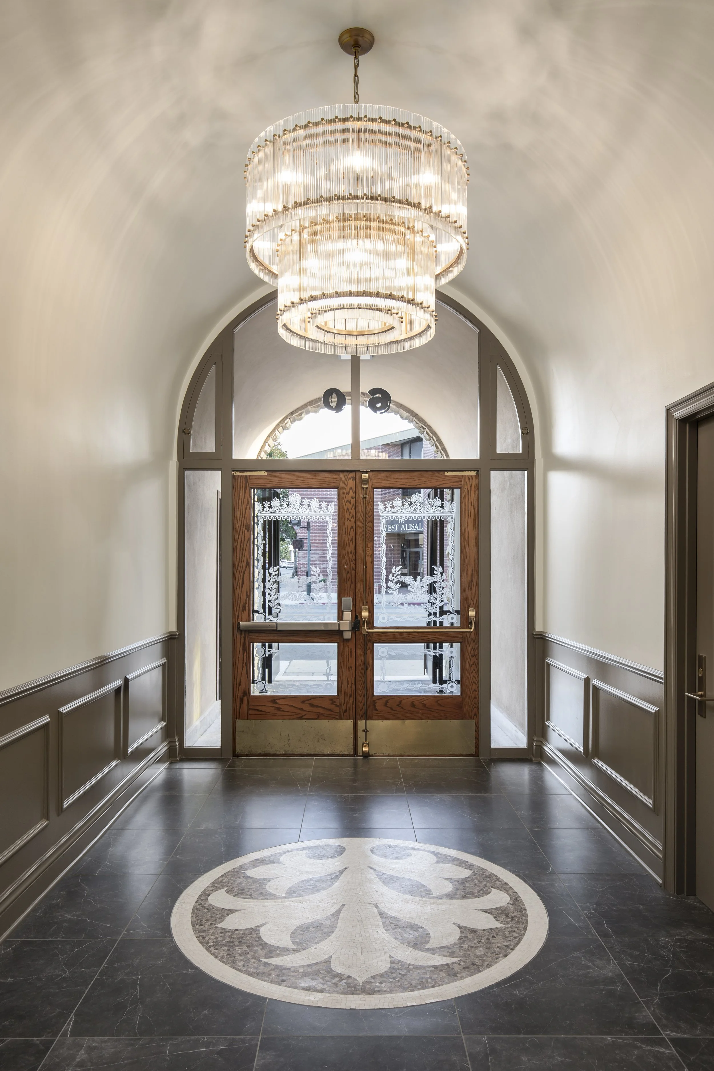 A grand entranceway with a large chandelier hanging from a vaulted ceiling. Double doors with etched glass panels lead outside, and a circular mosaic pattern adorns the floor.