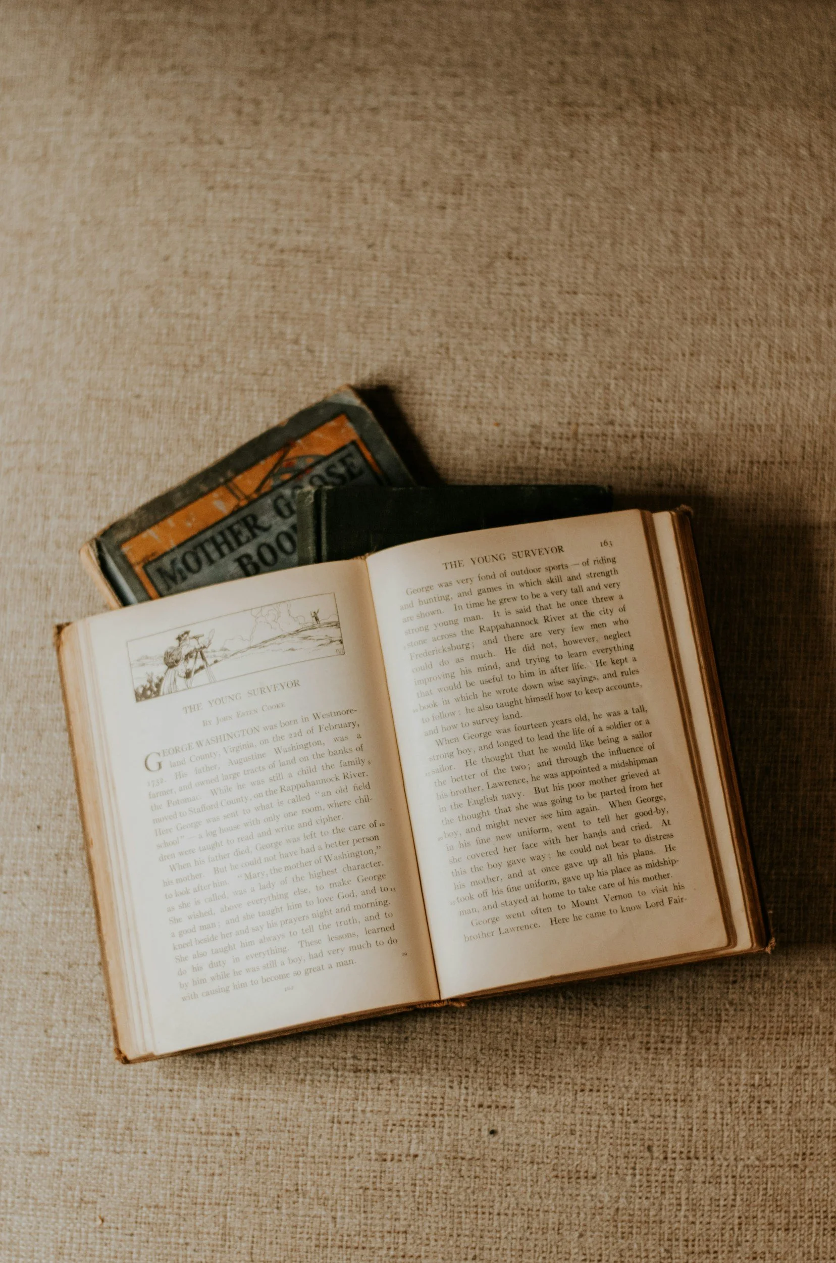 Open vintage book featuring a chapter about George Washington, with another old book partially visible on a textured fabric surface