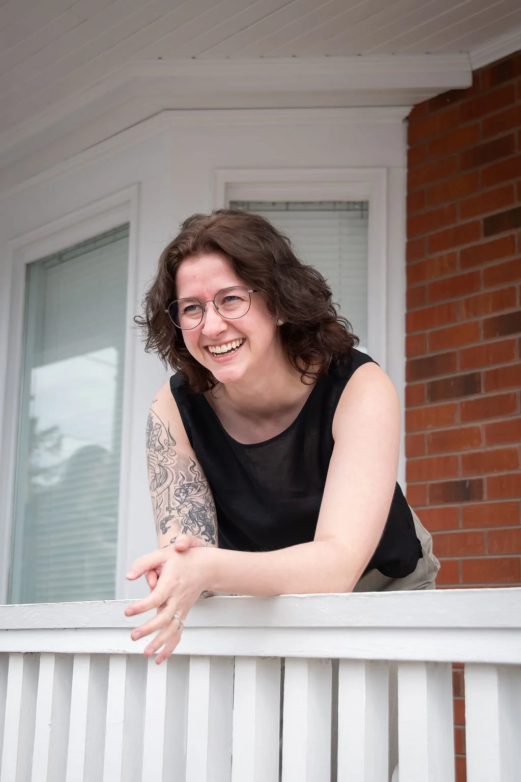 Smiling woman with glasses and a tattoo leaning on a white porch railing