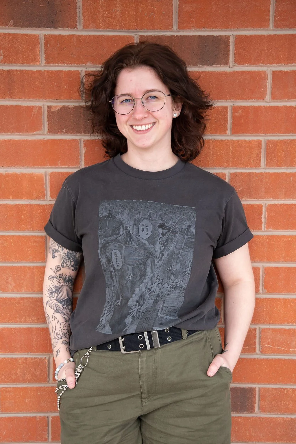 Smiling person wearing glasses, a graphic T-shirt, olive pants, and a belt while standing against a brick wall