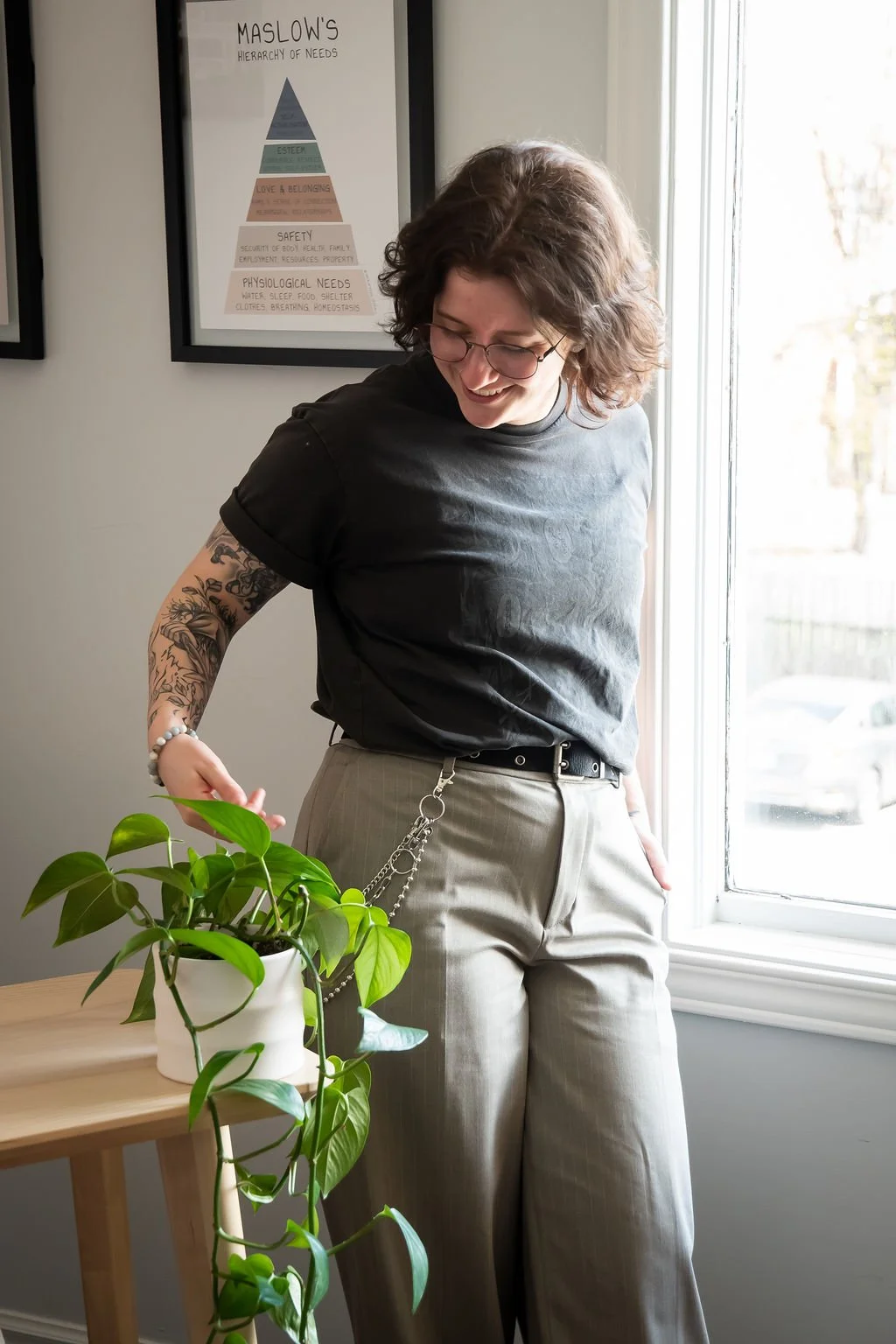 Smiling tattooed person in glasses tending to a potted trailing houseplant by a sunny window