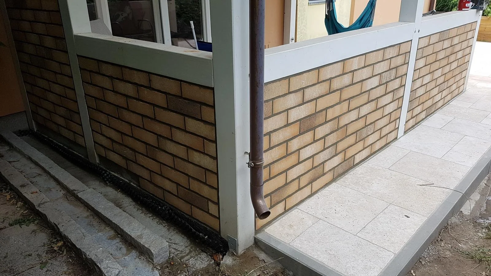 Eckansicht eines Außenbereichs mit gemauertem Wandschutz, weißen Rahmen und einem Regenfallrohr an der Ecke, der Boden ist mit Pflastersteinen ausgelegt.
