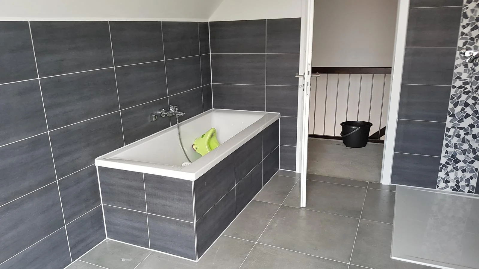 Projektentwicklung Berlin Ein moderner Badezimmer mit einer weißen Badewanne, dunklen Wandfliesen und einer Tür, die zu einem kleineren Raum führt, in dem ein schwarzer Eimer steht.