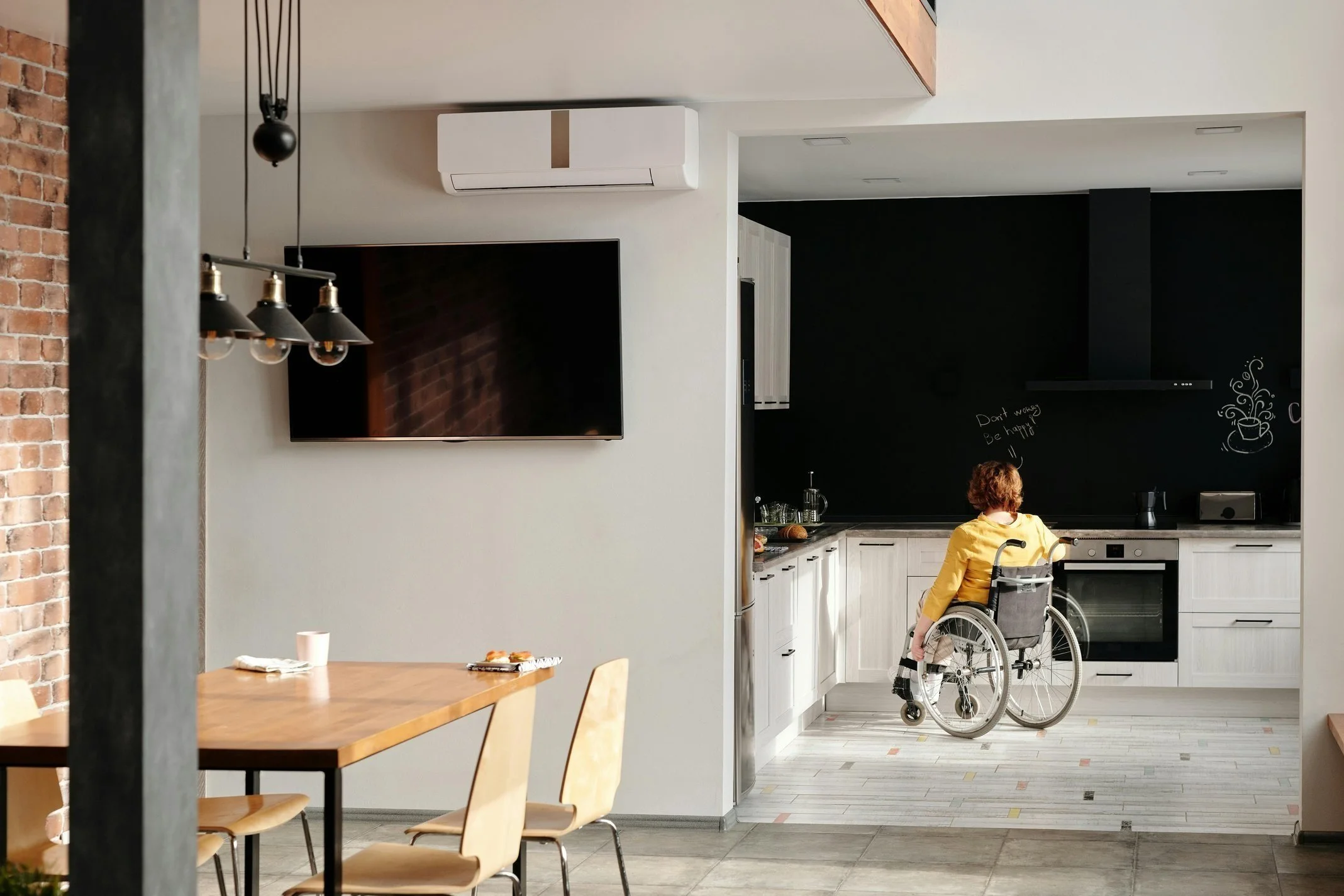 A person in a yellow shirt in a wheelchair is in a modern kitchen with white cabinets, a black wall with chalk writing, and a black range hood. The kitchen is connected to a dining area with a wooden table and beige chairs. A flat-screen TV is mounted on the wall, and there is a split air conditioner above it.