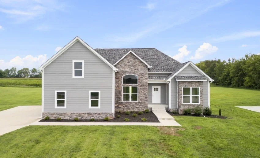 A modern suburban house with a gray exterior, stone accents, and a shingled roof, surrounded by a grassy lawn and trees in the background.