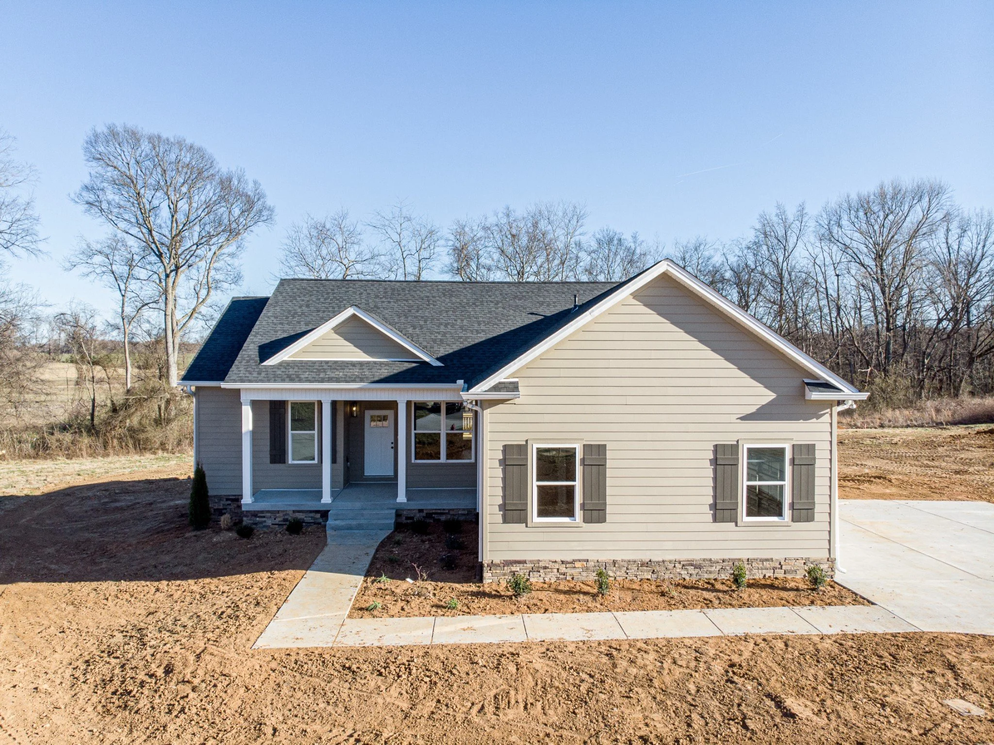 Modern single-story house with gray siding, black roof, and brick foundation, surrounded by bare ground and minimal landscaping, with trees and a clear sky in the background.