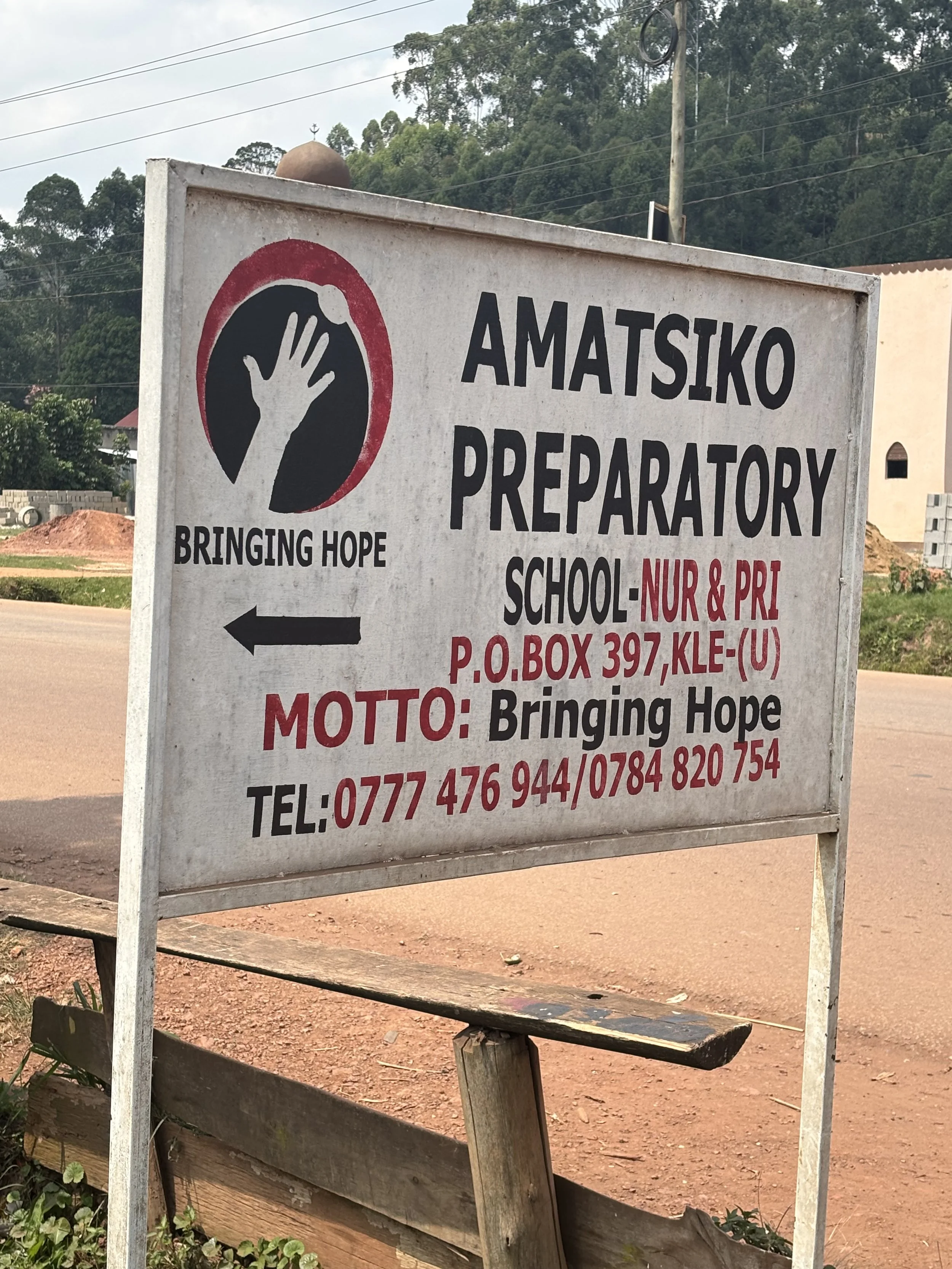 A sign for Amatsiko Preparatory School with the slogan "Bringing Hope" and contact information. The sign features a black hand symbol inside a red circle and a white background, located outdoors with trees and power lines in the background.