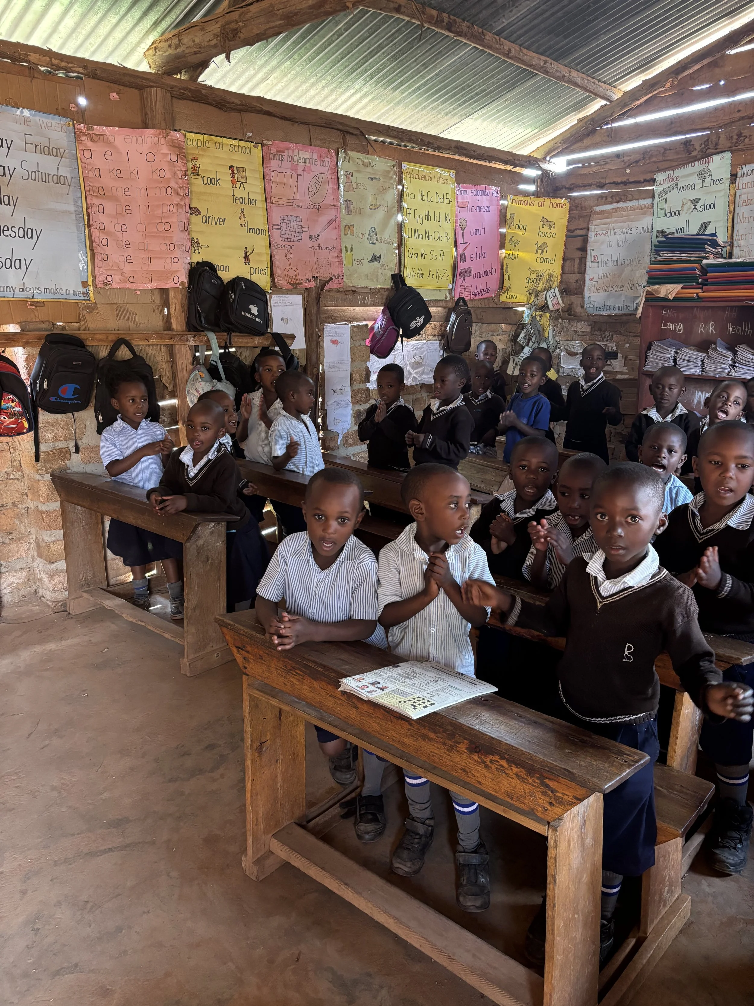 Group of young students inside a classroom with wooden benches and colorful educational posters on the wall, some students standing and others sitting, engaged and smiling.