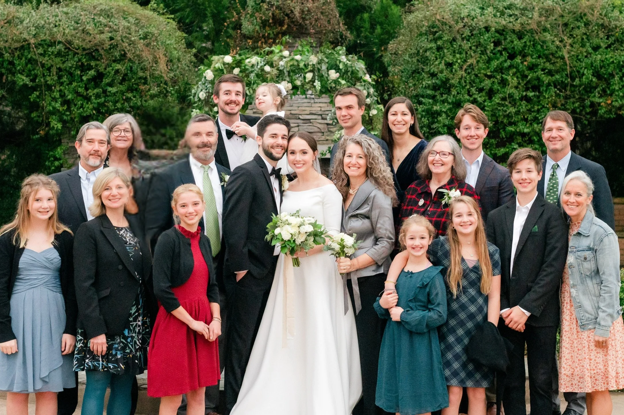 Group of people at a wedding, including the bride and groom, family, and children, standing outdoors in front of greenery and a floral backdrop.