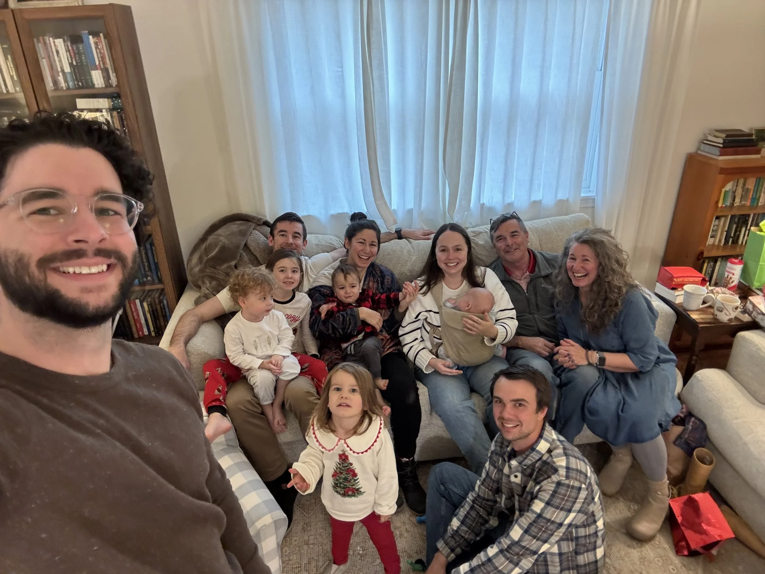 A group of ten people, including children and adults, gathered in a living room, smiling for a group photo with a man taking the selfie. There are bookshelves, curtains, and various household items visible in the background.