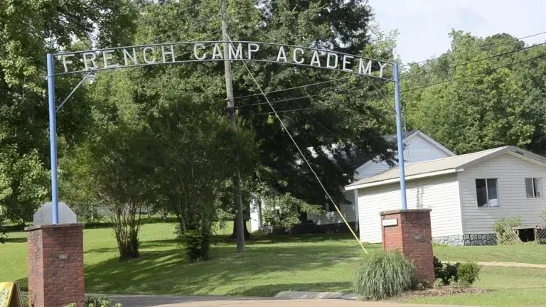 Entrance sign for French Camp Academy with brick pillars and a blue metal archway, surrounded by green trees and residential houses in the background.
