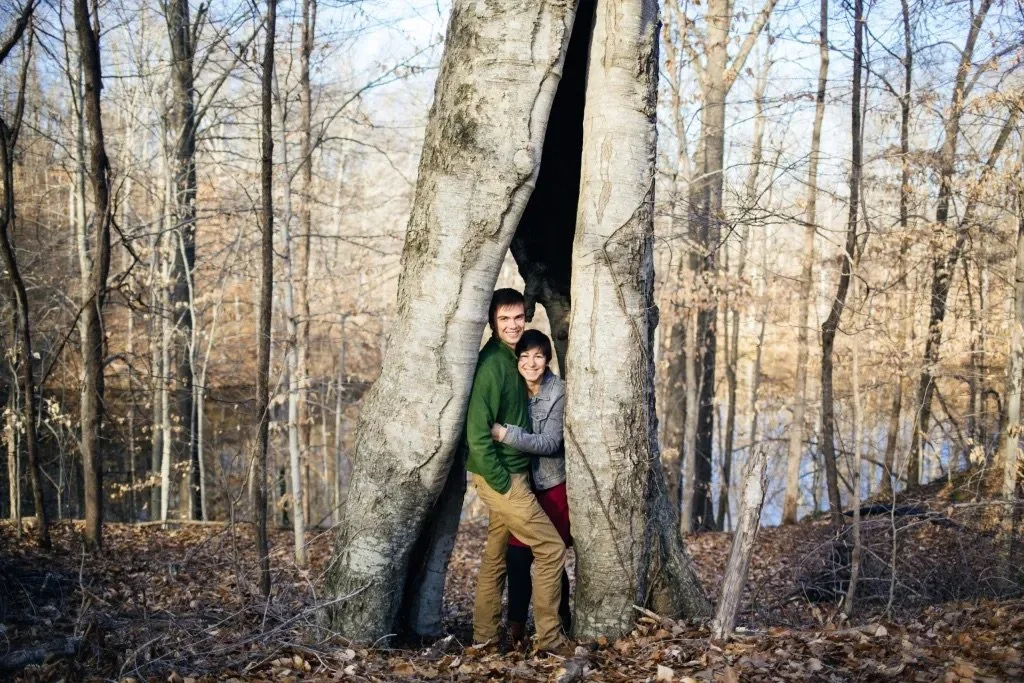 A couple standing inside the hollow of a large tree in a forest during autumn, with bare trees and fallen leaves all around.