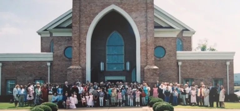 Large group of people gathered outside a brick church with a tall, arched entryway and stained glass windows during daytime.