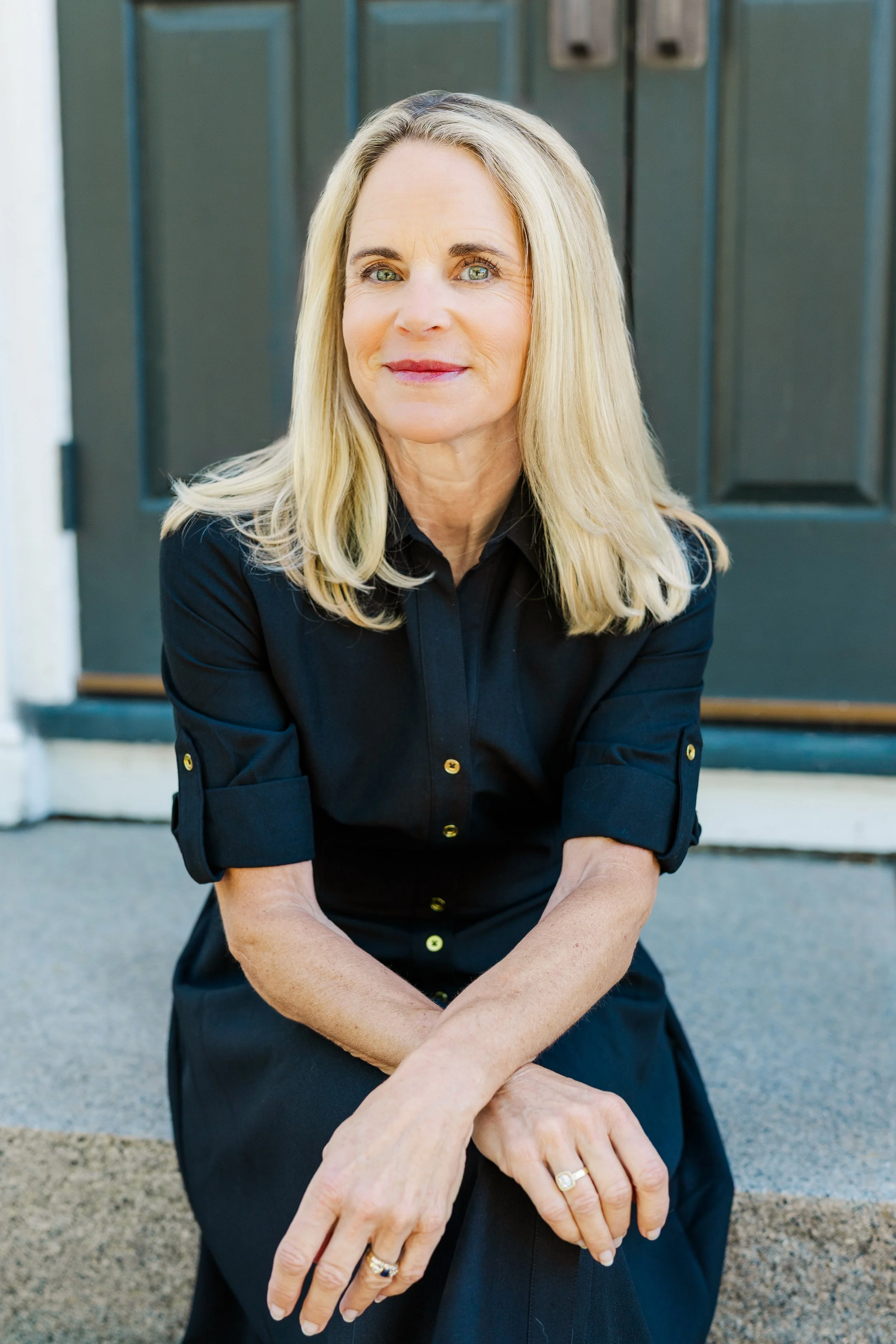 A middle-aged woman with blonde hair and blue eyes sitting on steps in front of a dark green door, wearing a black dress with gold buttons and rings on her fingers.