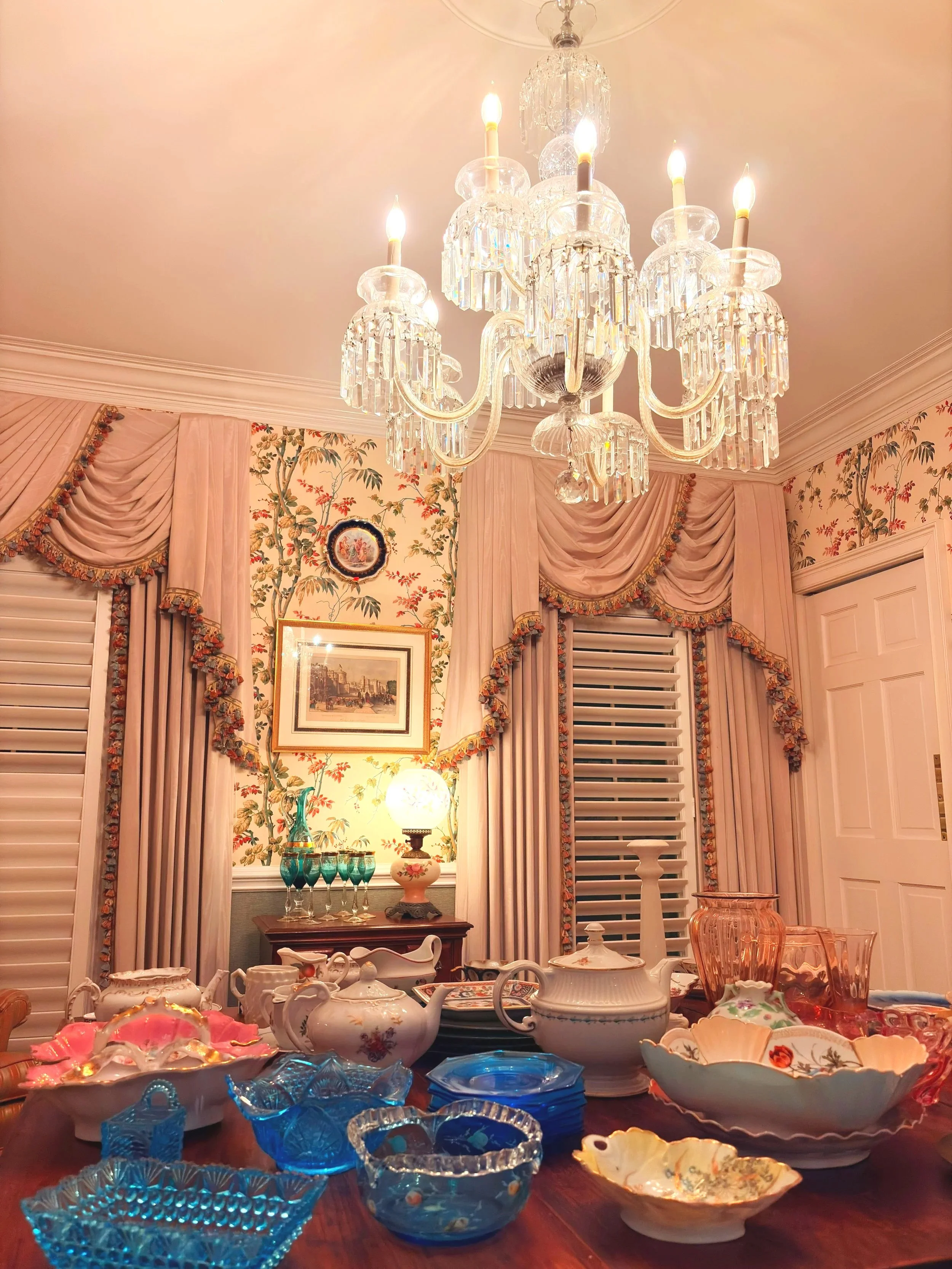 A chandelier hanging from a beige ceiling in a decorated room with pink drapes and floral wallpaper. The room has a table with various vintage glassware and porcelain dishes, including teapots, bowls, and glass cups in different colors like blue, pink, and clear.