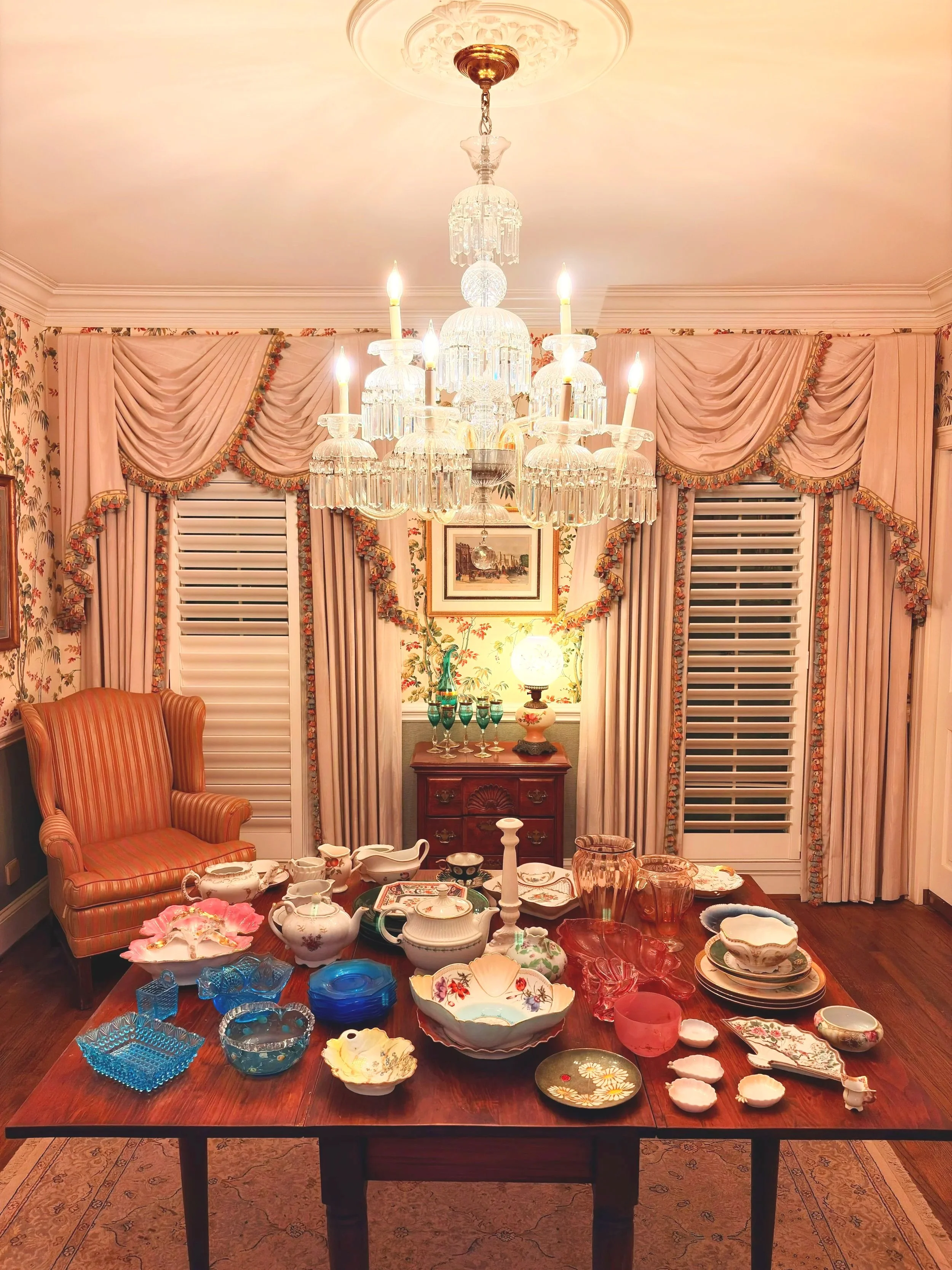A dining room with a large wooden table covered with various decorative glass and porcelain dishes and bowls. In the background, a crystal chandelier hangs from the ceiling, and floral patterned wallpaper and curtains frame the windows.