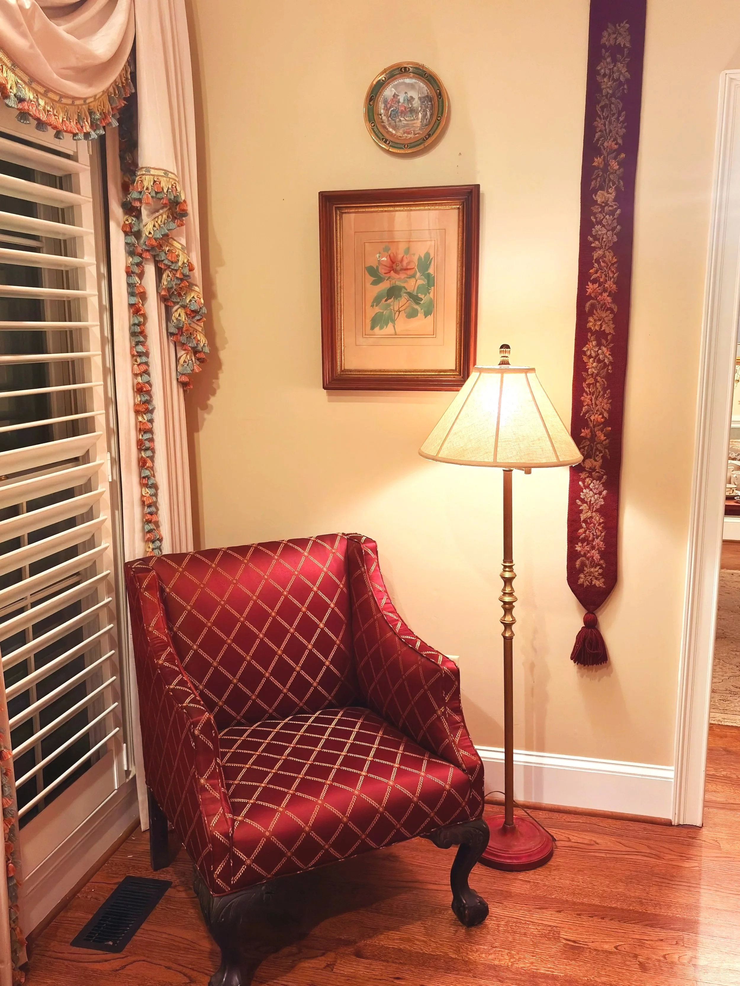 A cozy corner with a red armchair, a tall floor lamp, framed art on the wall, and decorative curtains.