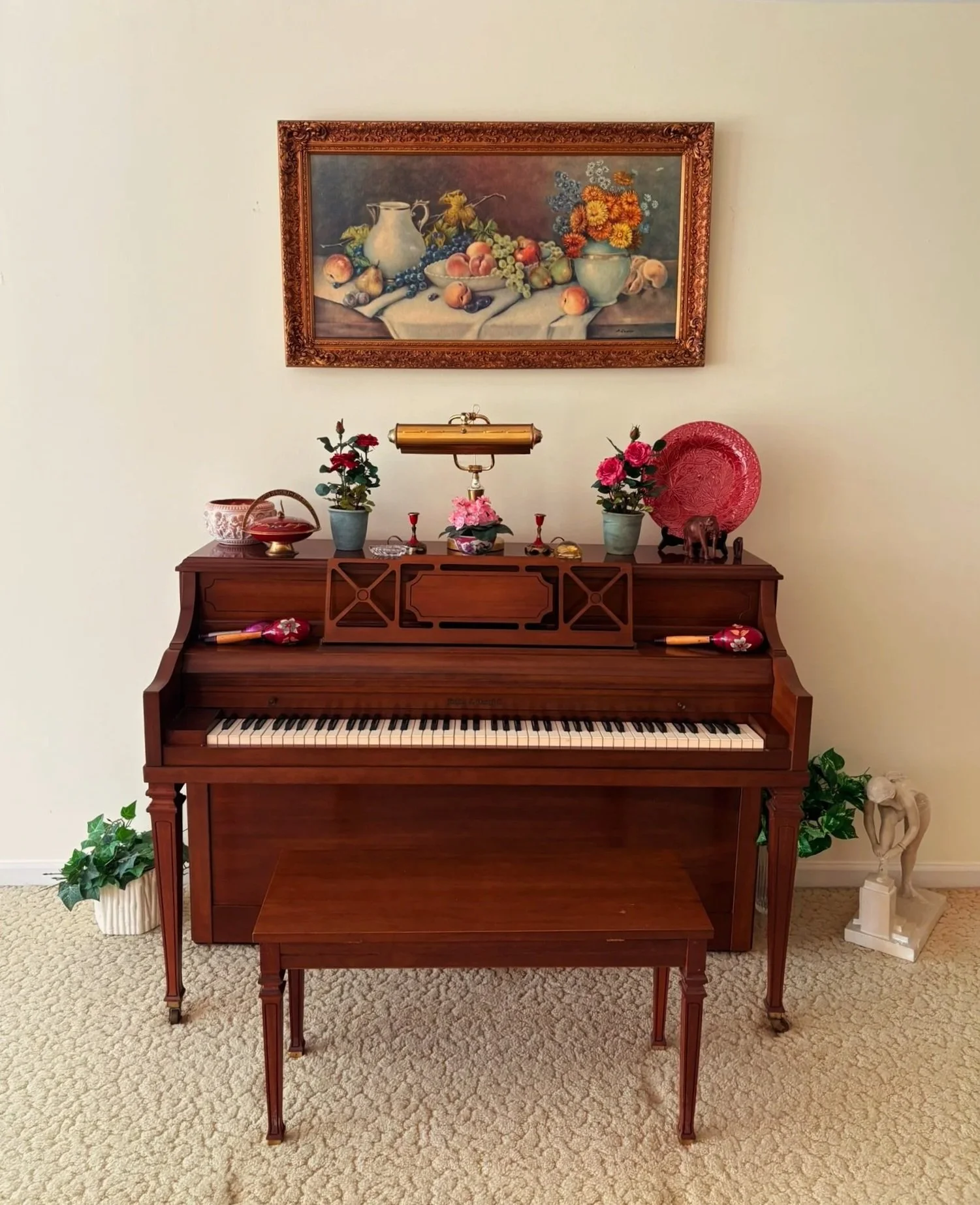 A wooden piano with a matching bench, decorated with potted flowers, a brass lamp, and various small decorative items, placed against a plain white wall with a framed still life painting of fruit and flowers hanging above.