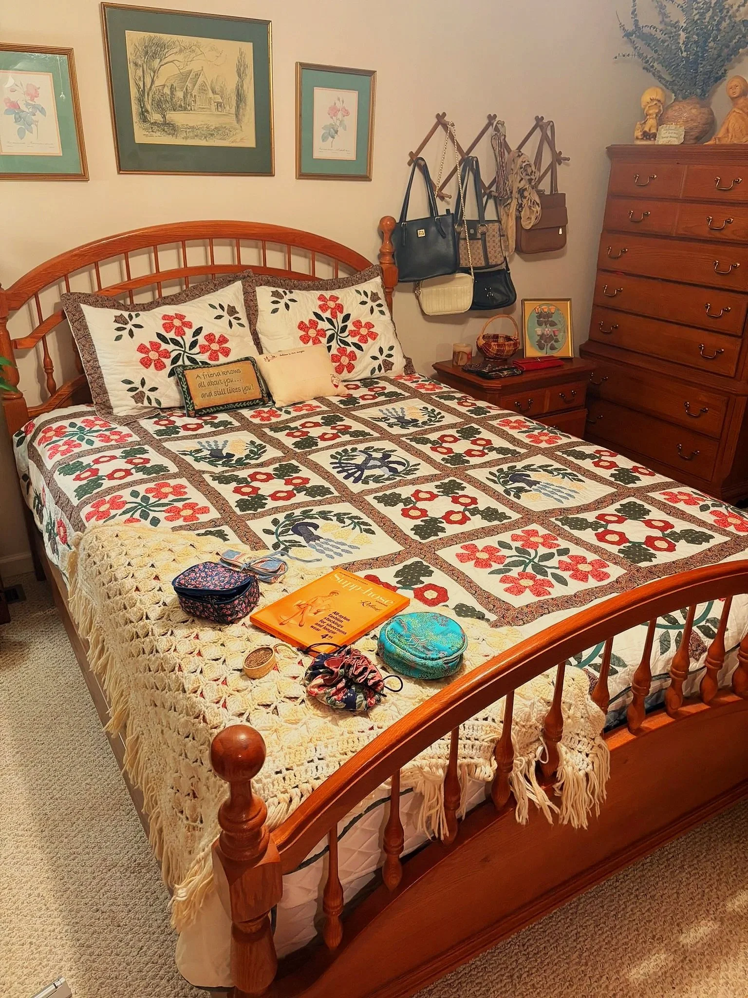 A bedroom with a wooden bed frame, floral quilt, decorative pillows, and a blanket. Items like books, a quilted bag, a patterned pouch, and a crochet piece are on the bed. A side table holds books, a framed picture, and small items. A wall has framed