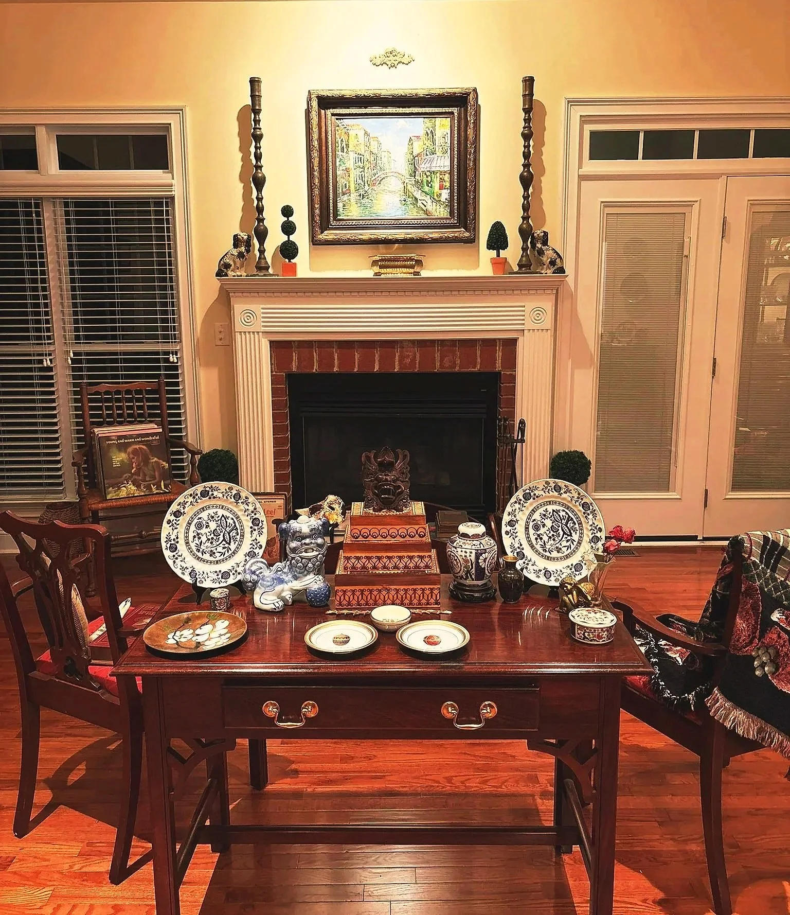 A cozy living room with a fireplace decorated with small potted plants, a framed painting of a city canal scene hangs above it, and a table set with decorative plates, vases, and figurines.