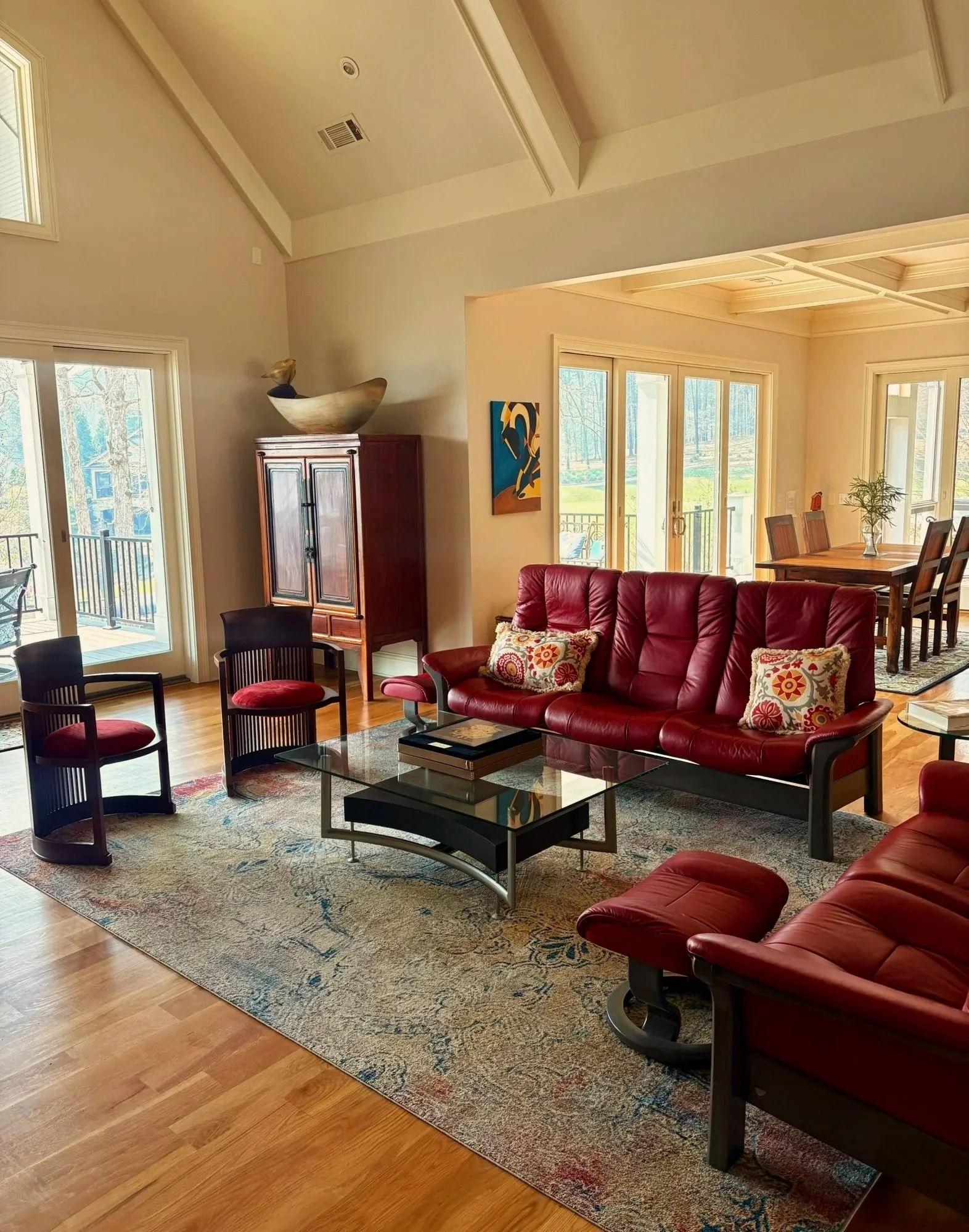 Living room with red leather furniture, a glass coffee table, a patterned rug, a wooden dining table with chairs, and large windows with outdoor views.