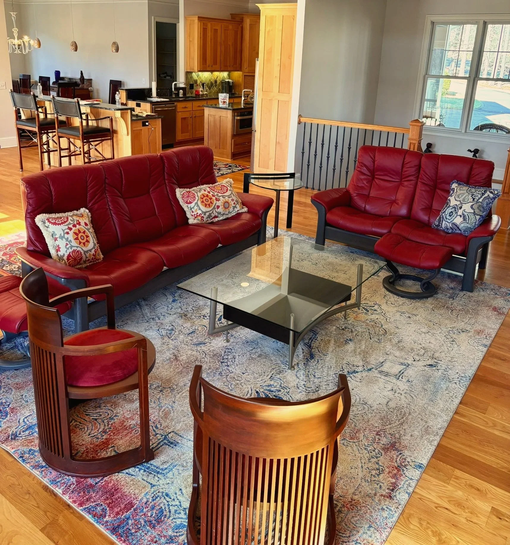 Living room with two red leather sofas, one with decorative pillows, a glass coffee table, and an area rug, open to a kitchen with wooden cabinets and a dining area with bar stools beneath pendant lights, hardwood floors, and large windows.