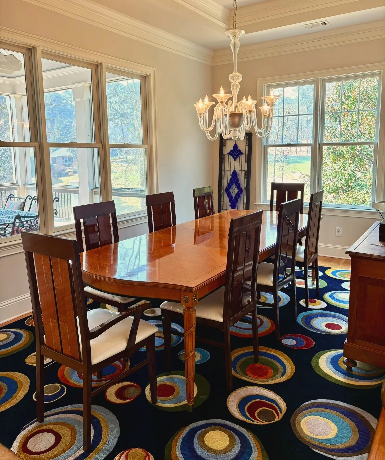 Dining room with a wooden table, eight chairs, a chandelier, large windows, and a colorful circular patterned carpet.