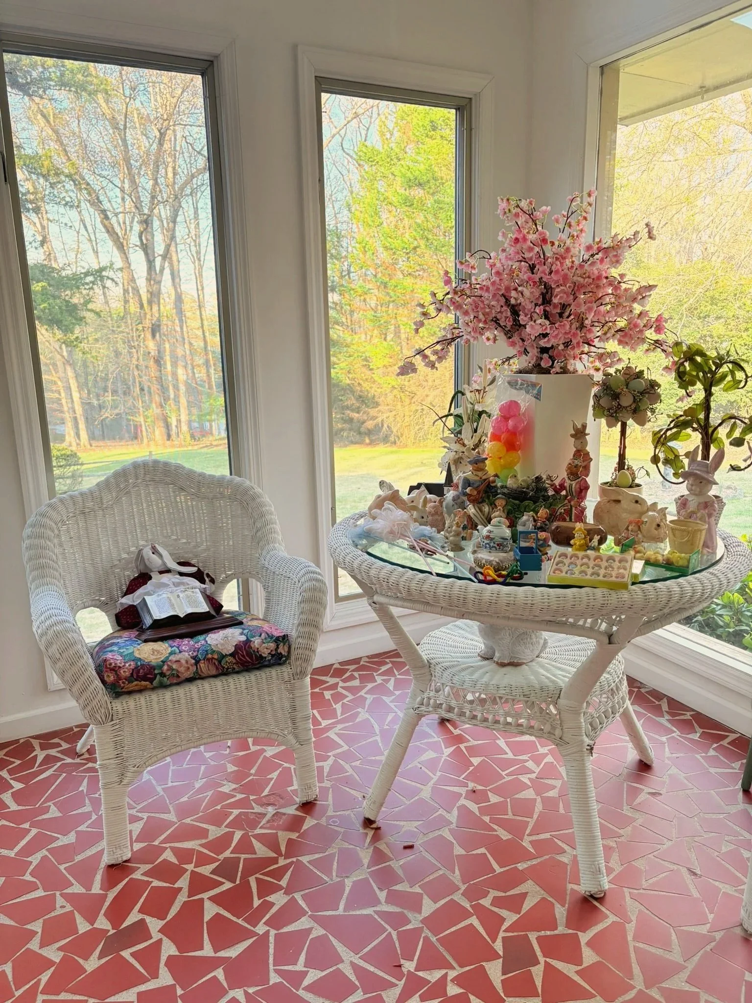 Sunlit indoor space with a round wicker table decorated with a large pink flower arrangement, surrounded by wicker chairs; window views of trees with green and yellow leaves, and a red and white irregular mosaic tiled floor.