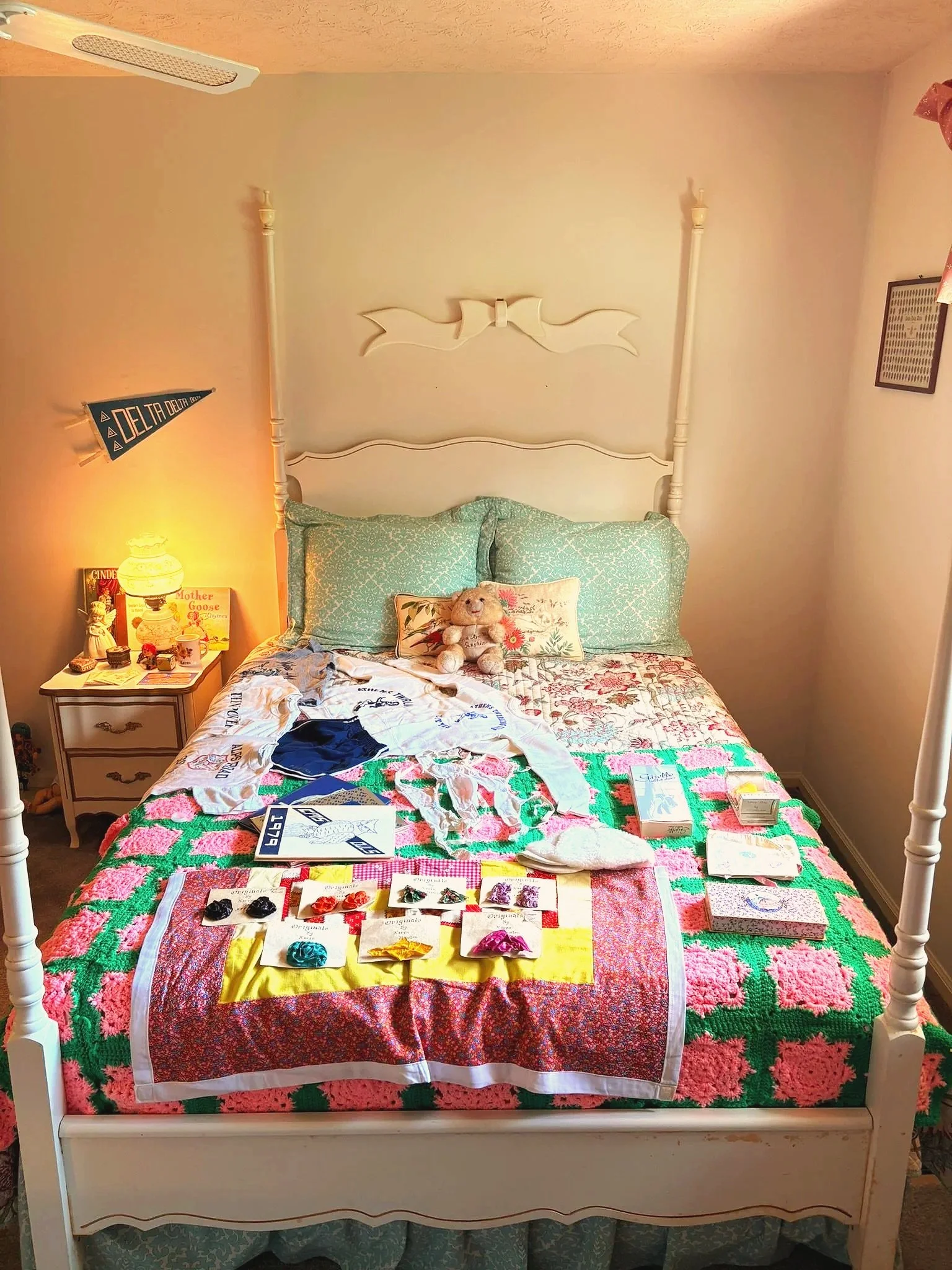 A bed with a white wooden headboard and four posted frame, covered with patchwork quilt, assorted pillows including a teddy bear, surrounded by various items including jewelry, books, and a yellow cloth on the mattress. A nightstand with decorative i