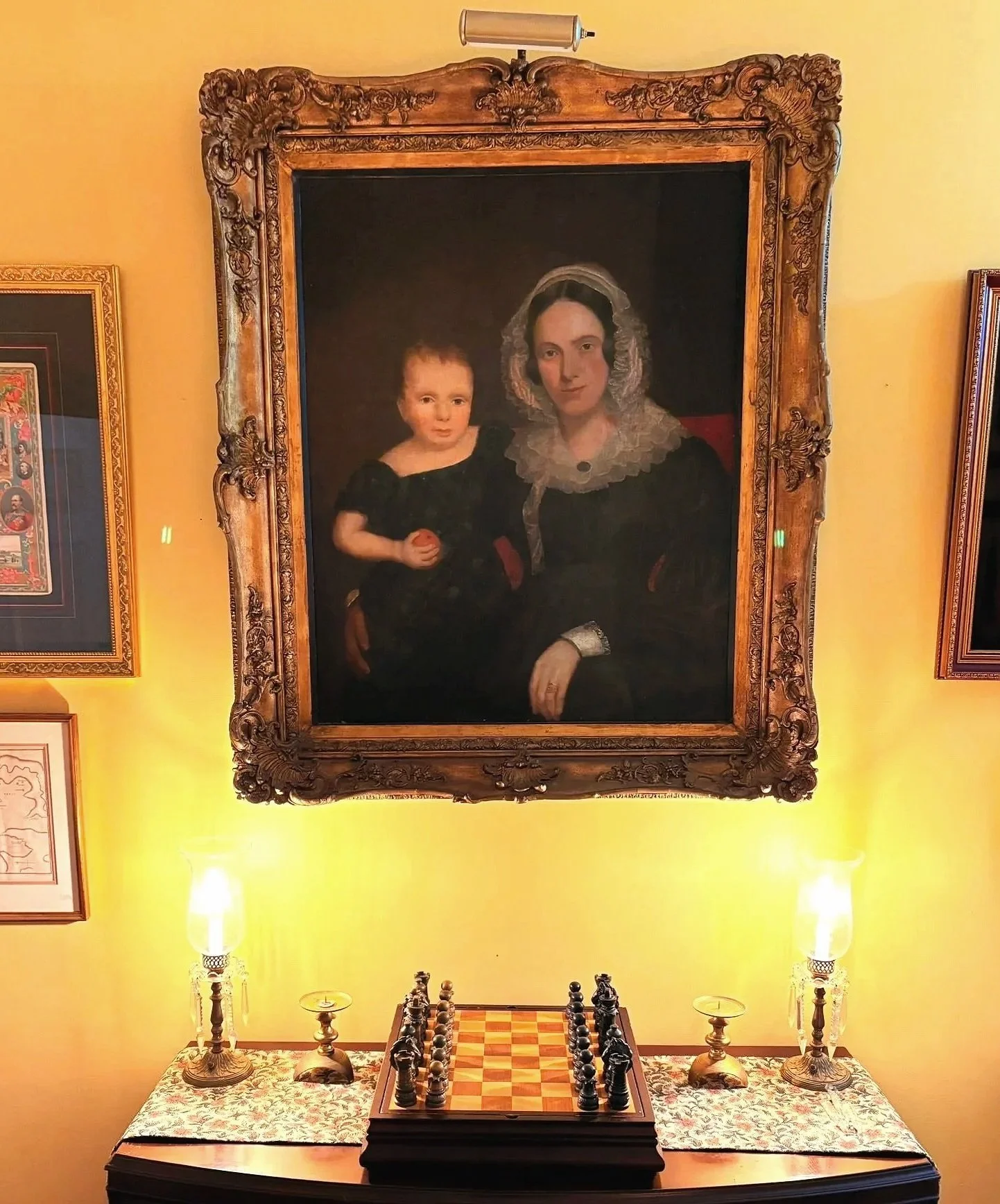 A large framed 19th century portrait of a woman with dark hair and a young child hanging on a yellow wall. Below the portrait, a chessboard with pieces is placed on a table covered with a patterned cloth, flanked by two ornate lamps.