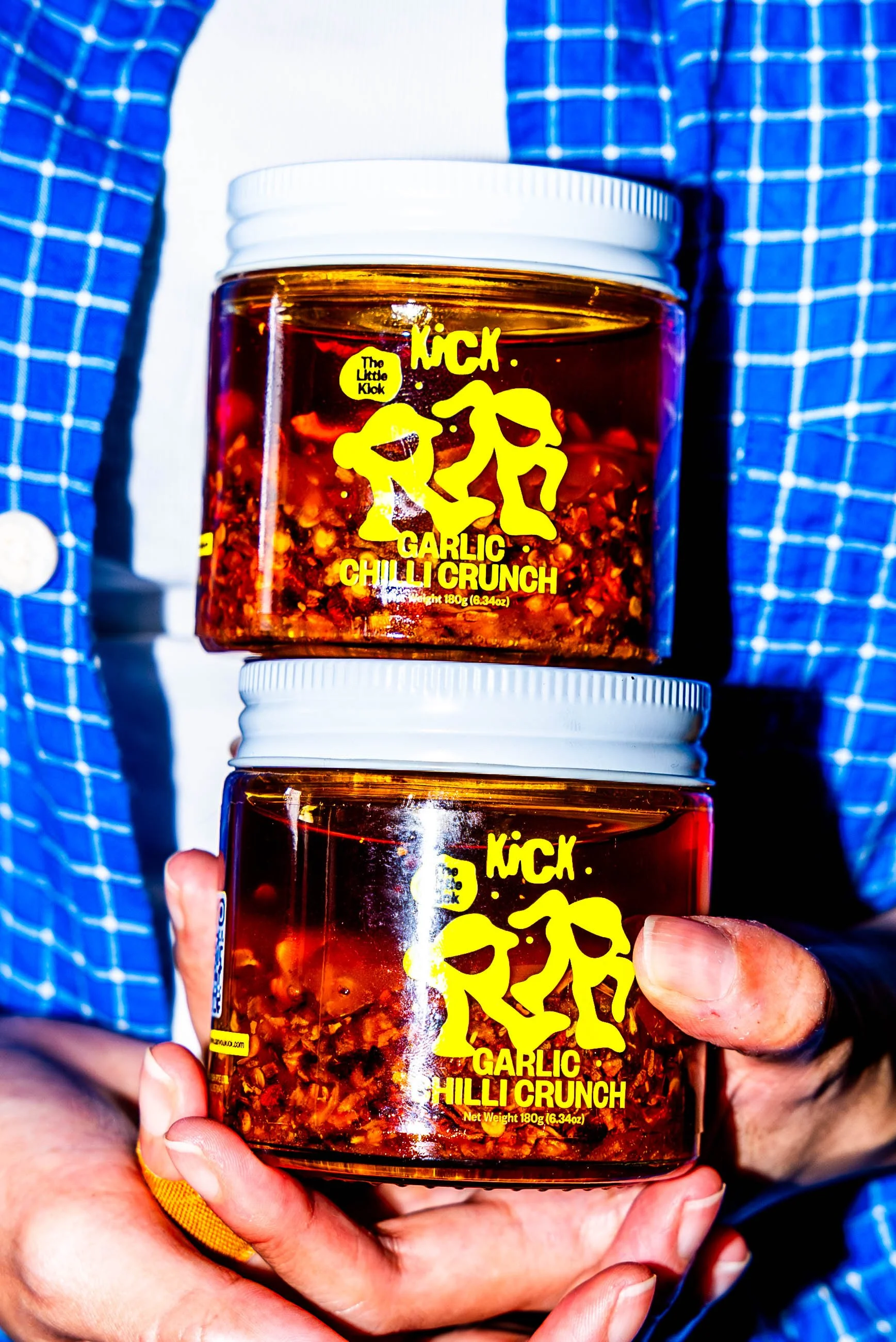 Two jars of Kick Garlic Chilli Crunch held by a person wearing a blue checkered shirt with a white background.