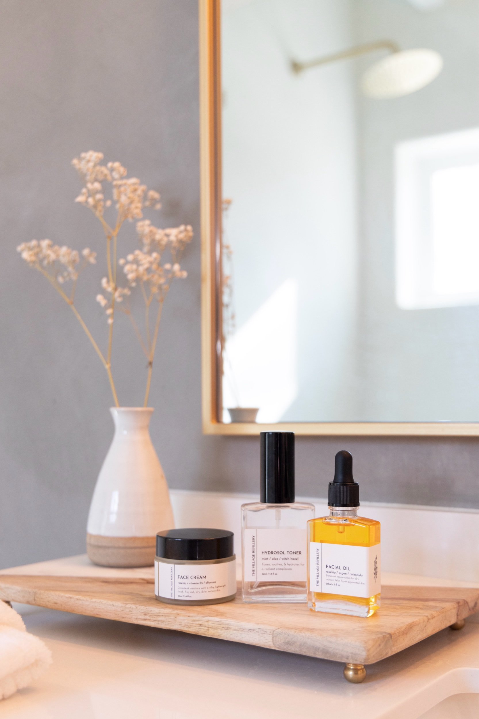 sink top display of a matte glass jar, glass atomizer spray bottle, and glass dropper bottle with white labels and black text reading "face cream" "hydrosol toner" and "face oil" respectively