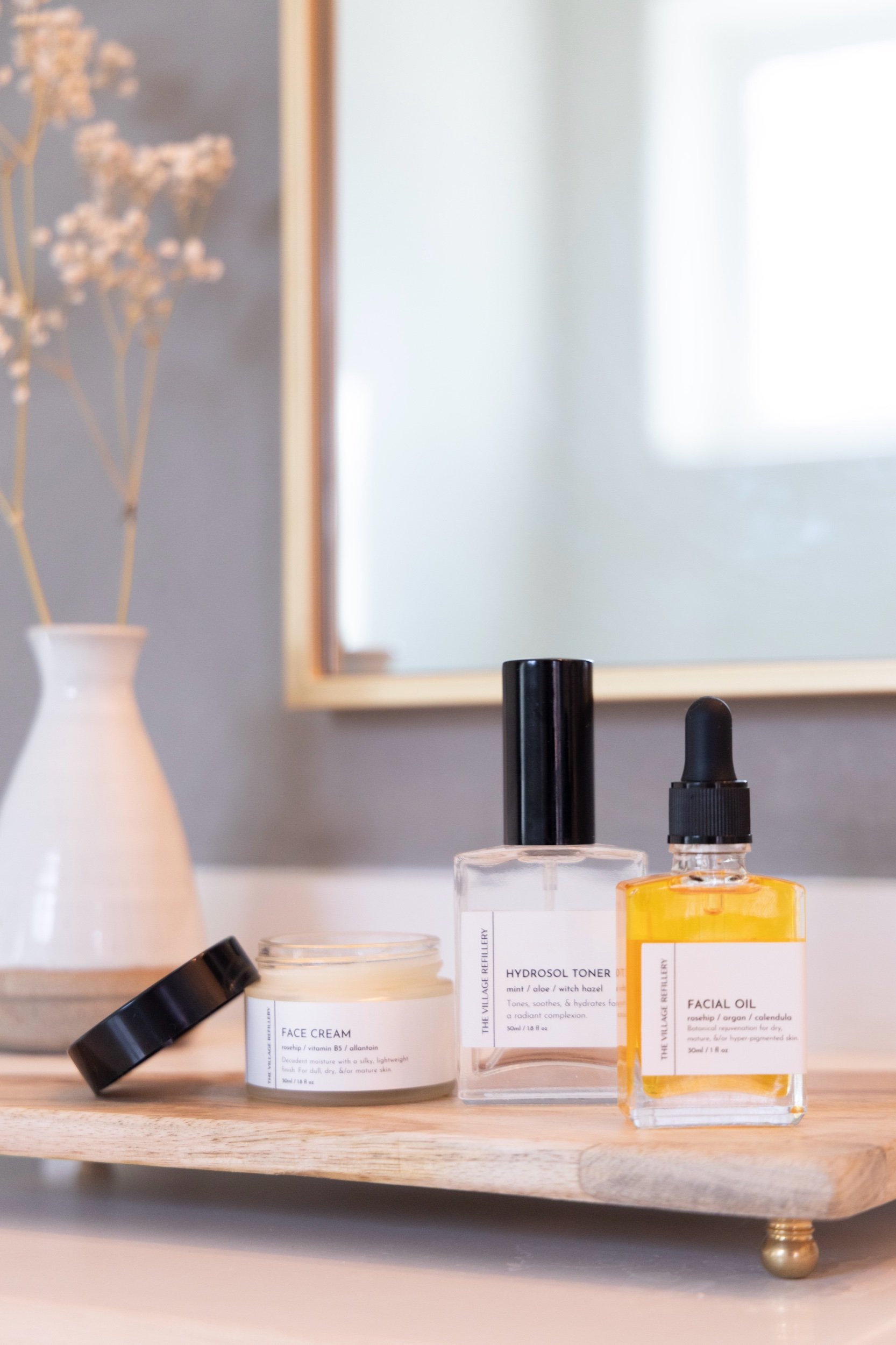 sink top display of a matte glass jar, glass atomizer spray bottle, and glass dropper bottle with white labels and black text reading "face cream" "hydrosol toner" and "face oil" respectively