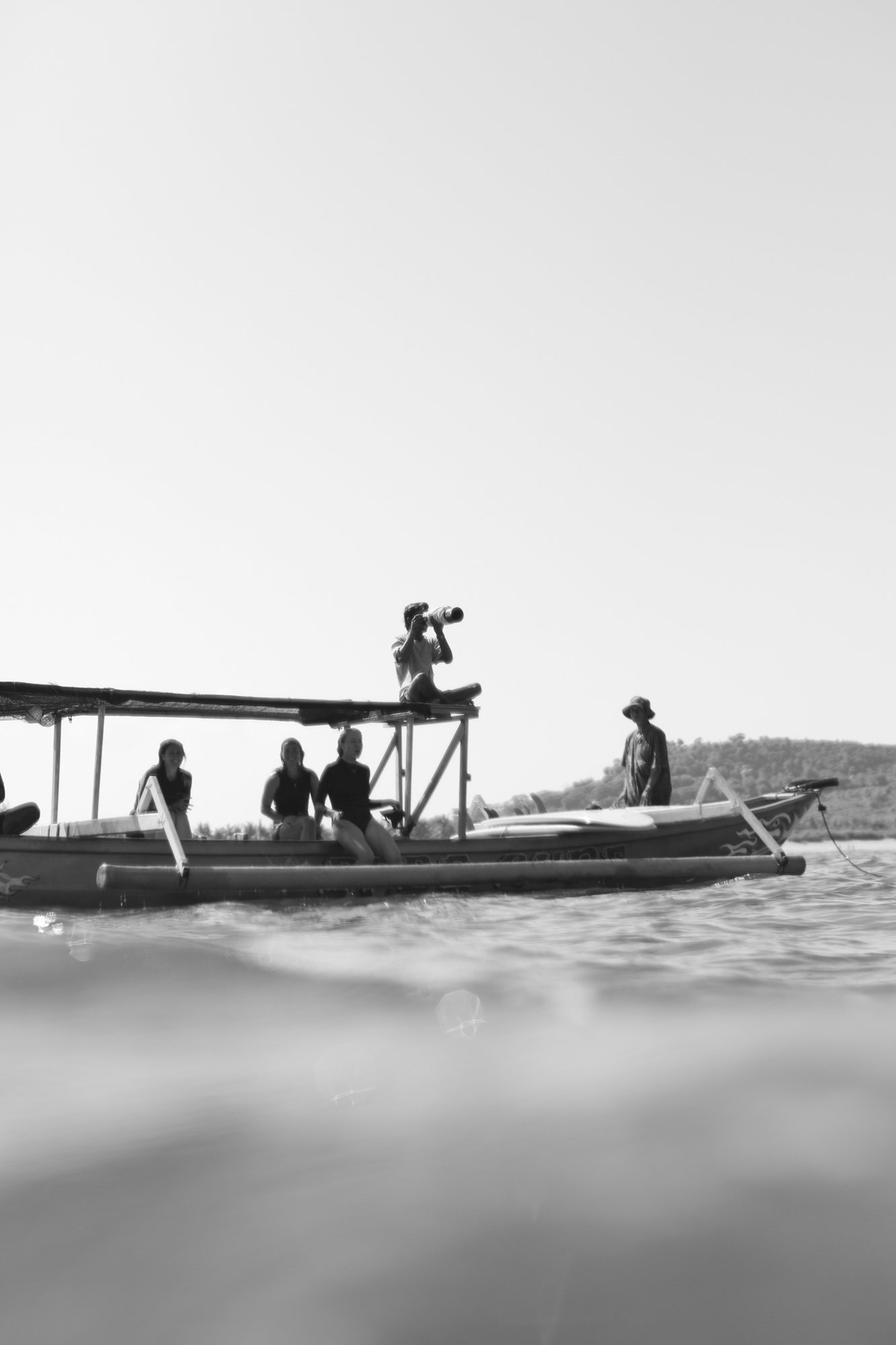 People sitting and standing on a boat on the water, with one person sitting on a platform with a camera, and a person standing on the boat, in a scenic outdoor setting.