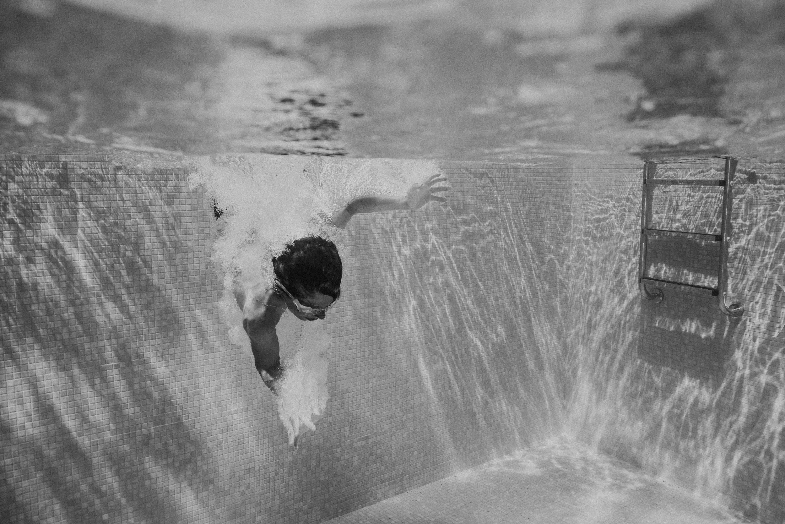 A person swimming underwater in a pool, holding a towel or cloth, wearing goggles, with pool ladder visible on the wall.